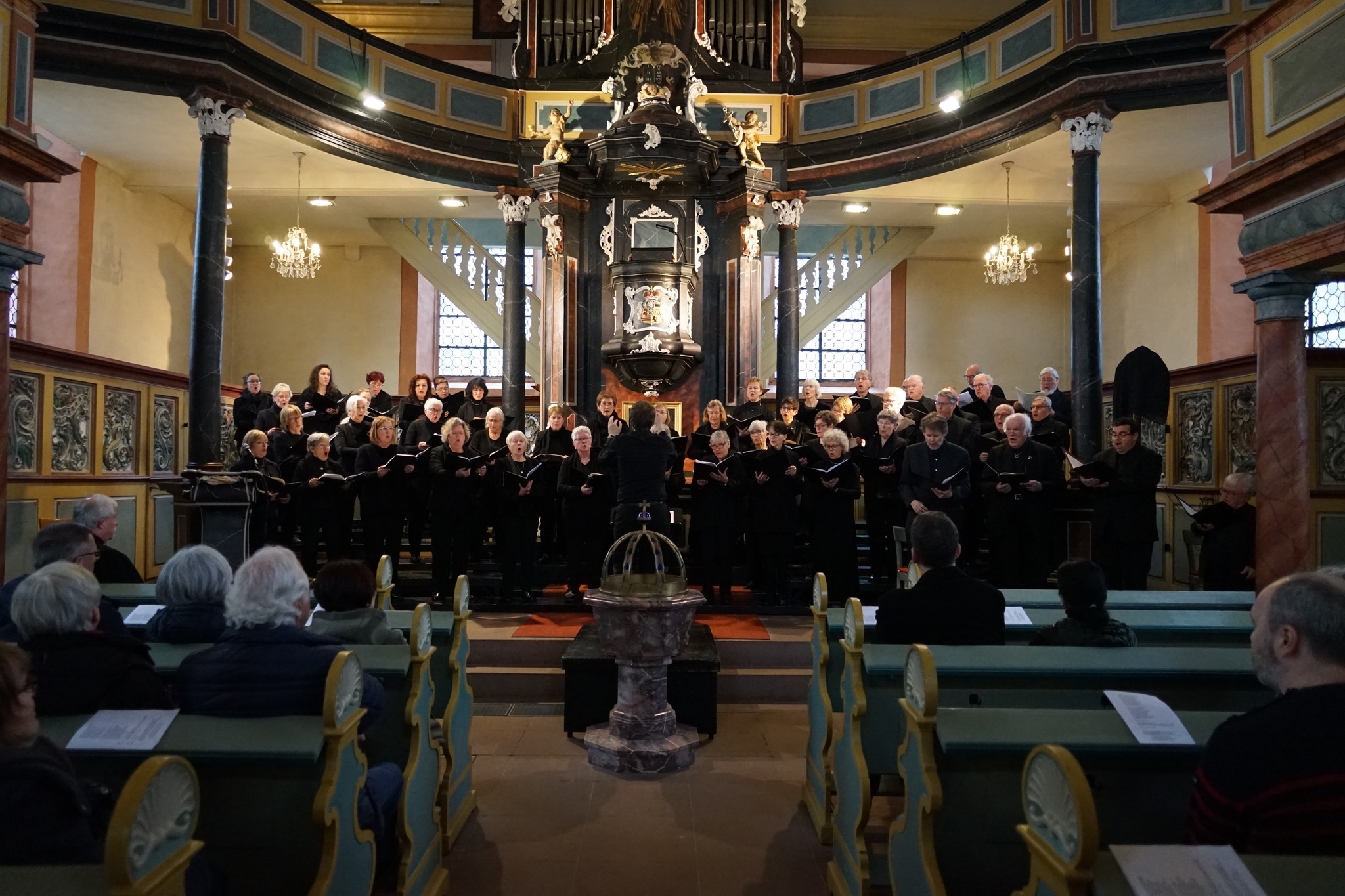 Ein gemischter Chor, 46 Personen, in schwarz gekleidet und mit schwarzen Gesangsbüchern in der Hand, steht im Altarraum. Hinter ihnen ein Hochaltar, dahinter zwei große Sprossenfenster und auf einer Empore die Orgel. Vorn sind Kirchenbänke mit Zuhörern zu sehen. Die Kirche ist in hell- und dunkelgrün und gold gehalten.