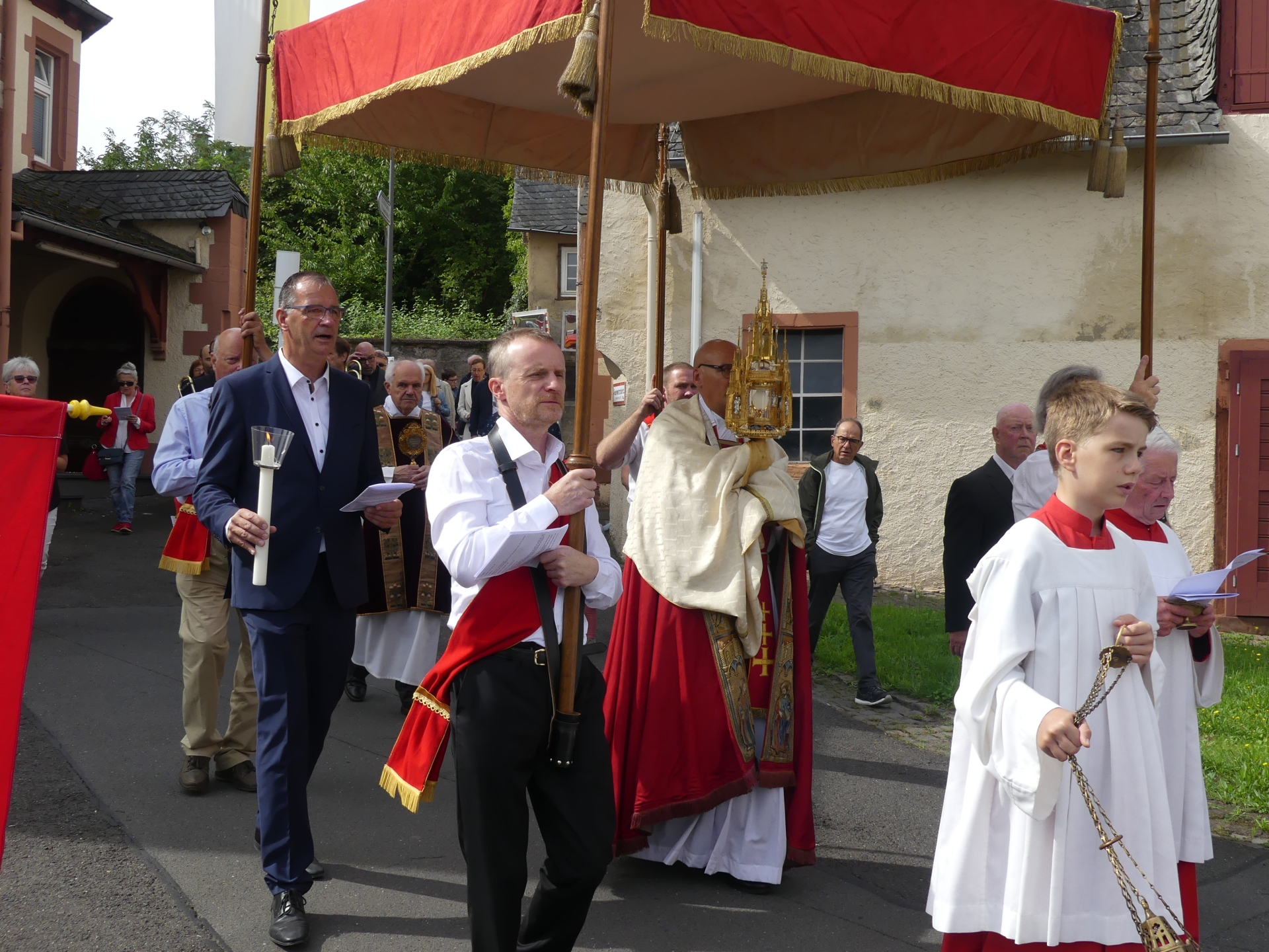 Unter einem roten Stoffdach mit goldenen Quasten, welches von vier Trägern an Holzstangen getragen wird, läuft der Pfarrer in rot-beige gekleider mit einem goldenen Gefäß mit einer Relique. Vorne schwänkt ein Messdiener in rot-weiß einen goldenen Weihrauchbehälter. Die Prozession wird von mehreren Personen begleitet.