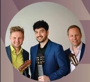 Drei Männer im Hemd und Sakko mit Ihren Gitarren in der Hand. Man sieht nur den Oberkörper. Links der Mann mit blonden Haaren und Bart trägt ein grünes Hemd und ein blaues Sakko, der Mittlere mit schwarzen Haaren und Bart ein blaues Sakko und ein schwarzes Hemd und der Rechte mit blonden Haaren und Bart ein weißes Hemd und schwarzes Sakko.