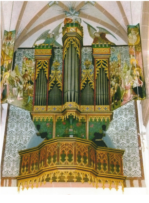 Eine Orgel mit mehreren Orgelpfeifen, die in fünf Rechtecken angeordnet sind, die mit goldenene Ornamenten verziert sind. Das Emporengeländer ist gold, grün und orange-braun dargestellt mit Ornamenten. HInter der Orgel sind auf weißem Untegrund graue Ornamente und neben und oberhalb der Orgelpfeifen sind Personen und Engel gemalt.