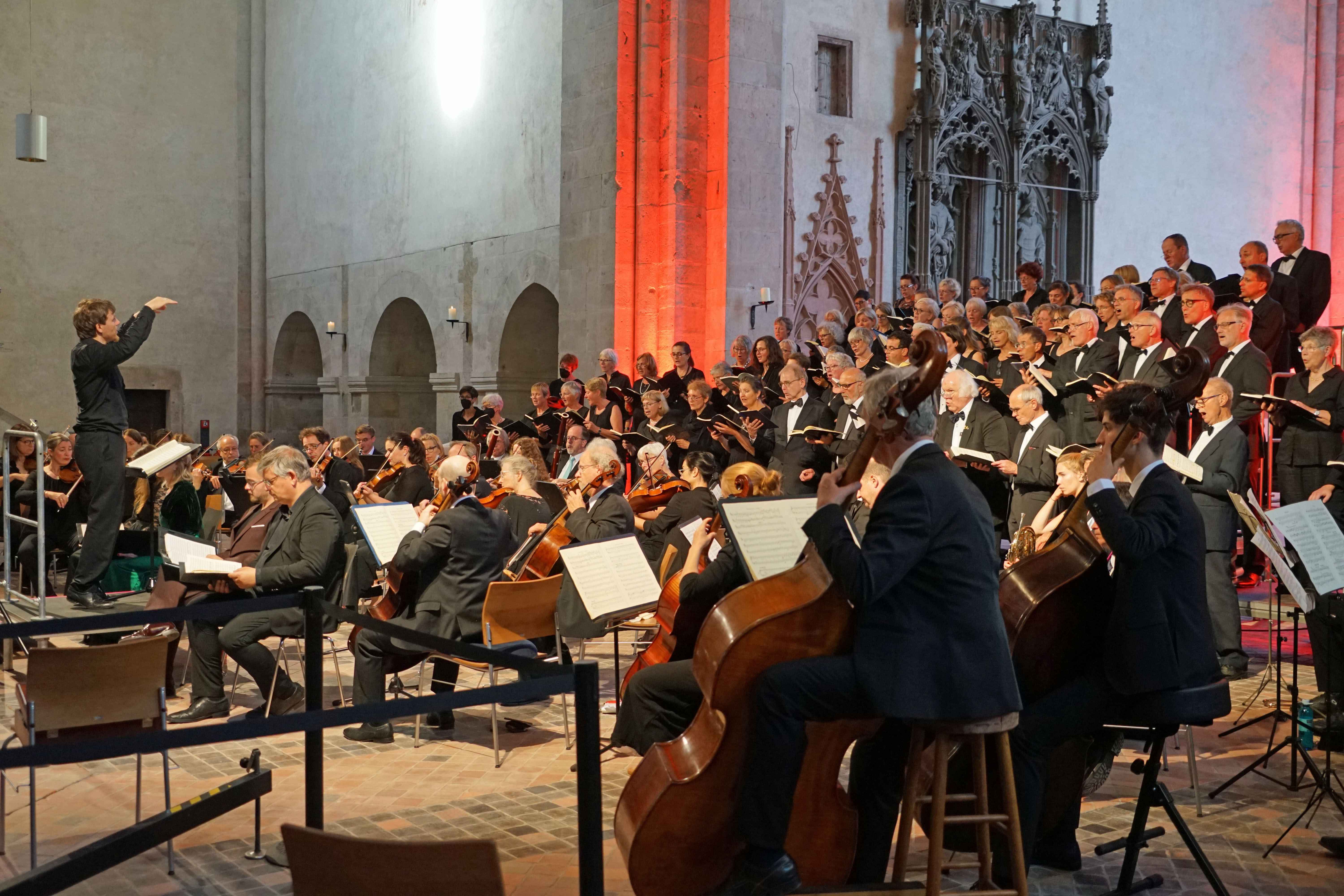 Ein großes Orchester sitzt vor einem großem Chor, der steht. Alle Männer haben schwarze Anzüge und weiße Hemden an. Die Frauen sind ebenso dunkel gekleidet. Im Vordergrund steht ein Dirigent auf einem Podest. Im Hintergrund ist eine Ecke rot angestrahlt und es sind drei Rundbögen in einer grauen Wand zu sehen. Ebenso ist ein rötliches und zwei grau stark verzierte Elemente aus Stein an der einen Wand.