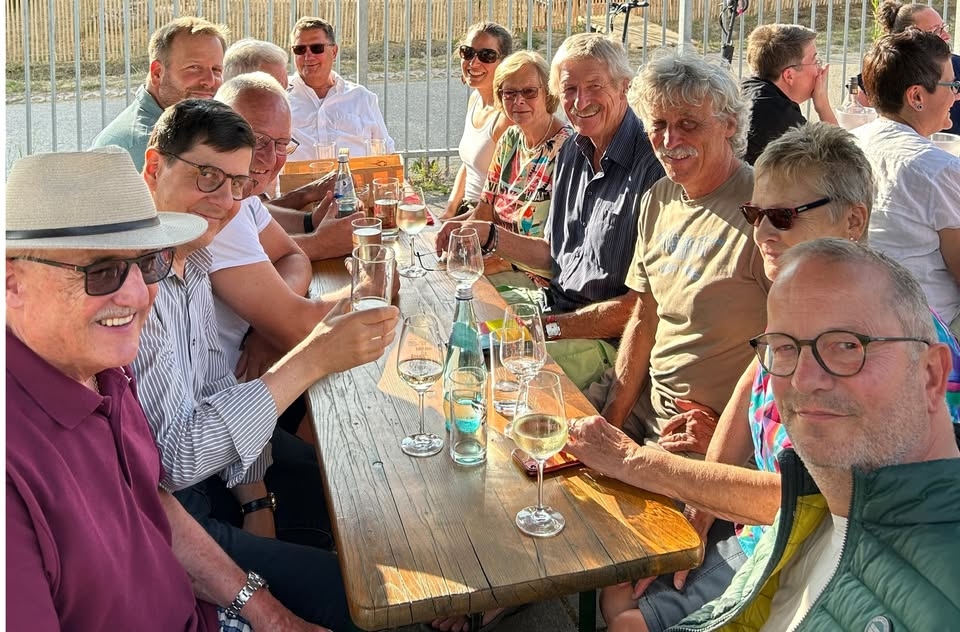 Auf einer Bierzeltgarnitur sitzen vier Frauen und acht Männer. Auf dem Tisch stehen gefüllte Wein- und Wassergläser und Wasserflaschen. Im Hintergrund ist ein Zaun aus Gitterstäben aus Metall und nach der Straße ein Zaun aus Holzstäben.