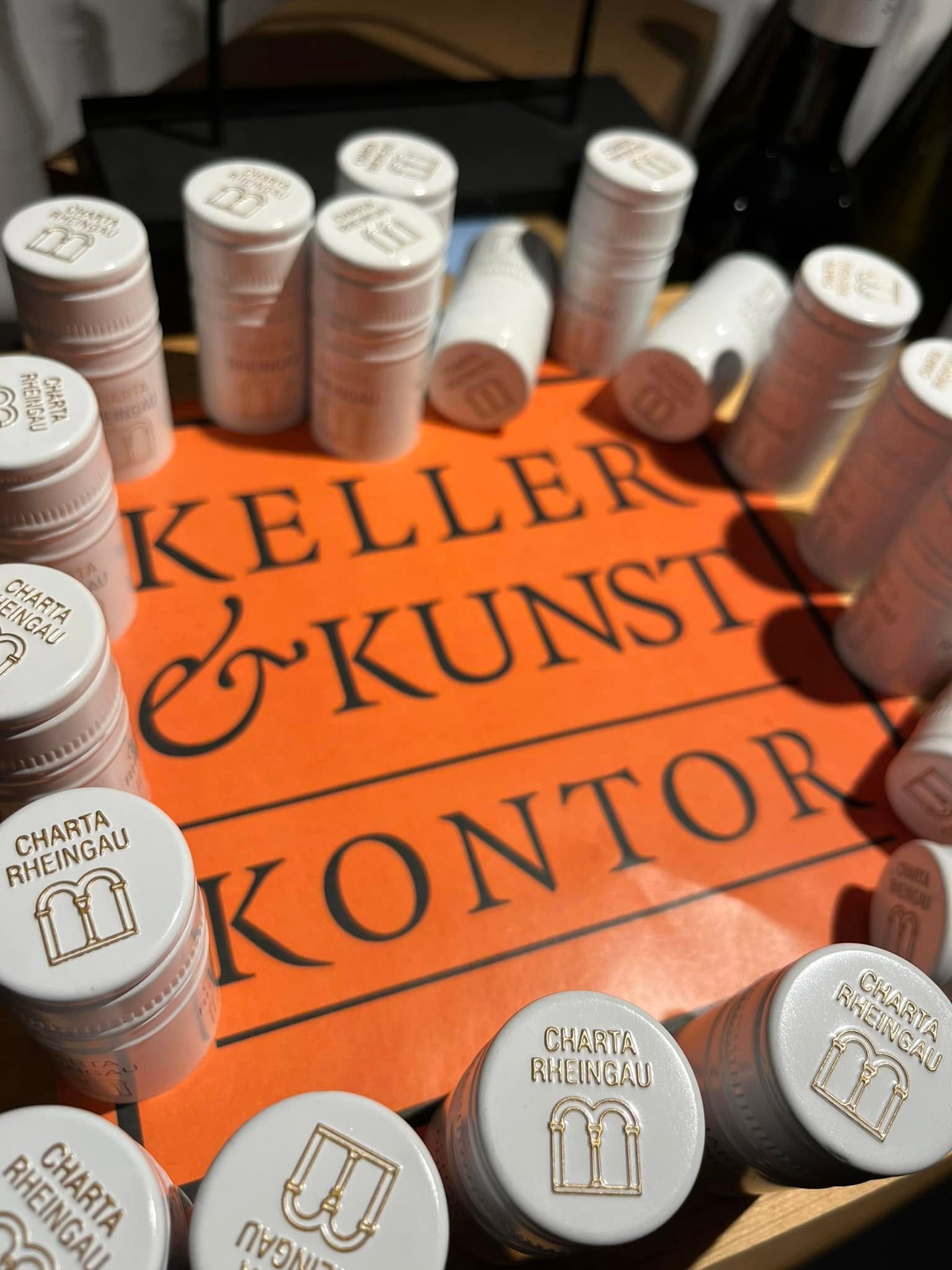 Auf einem orangenem Quadrat steht in schwarten Buchstaben Keller & Kunst Kontor. Drum herum sind weiße Aluschrauber für Weinflaschen, auf denen in goldener Schrift Charta Rheingau mit zwei Fensterbögen abgebildet ist. Vier Hülsen sind umgefallen.