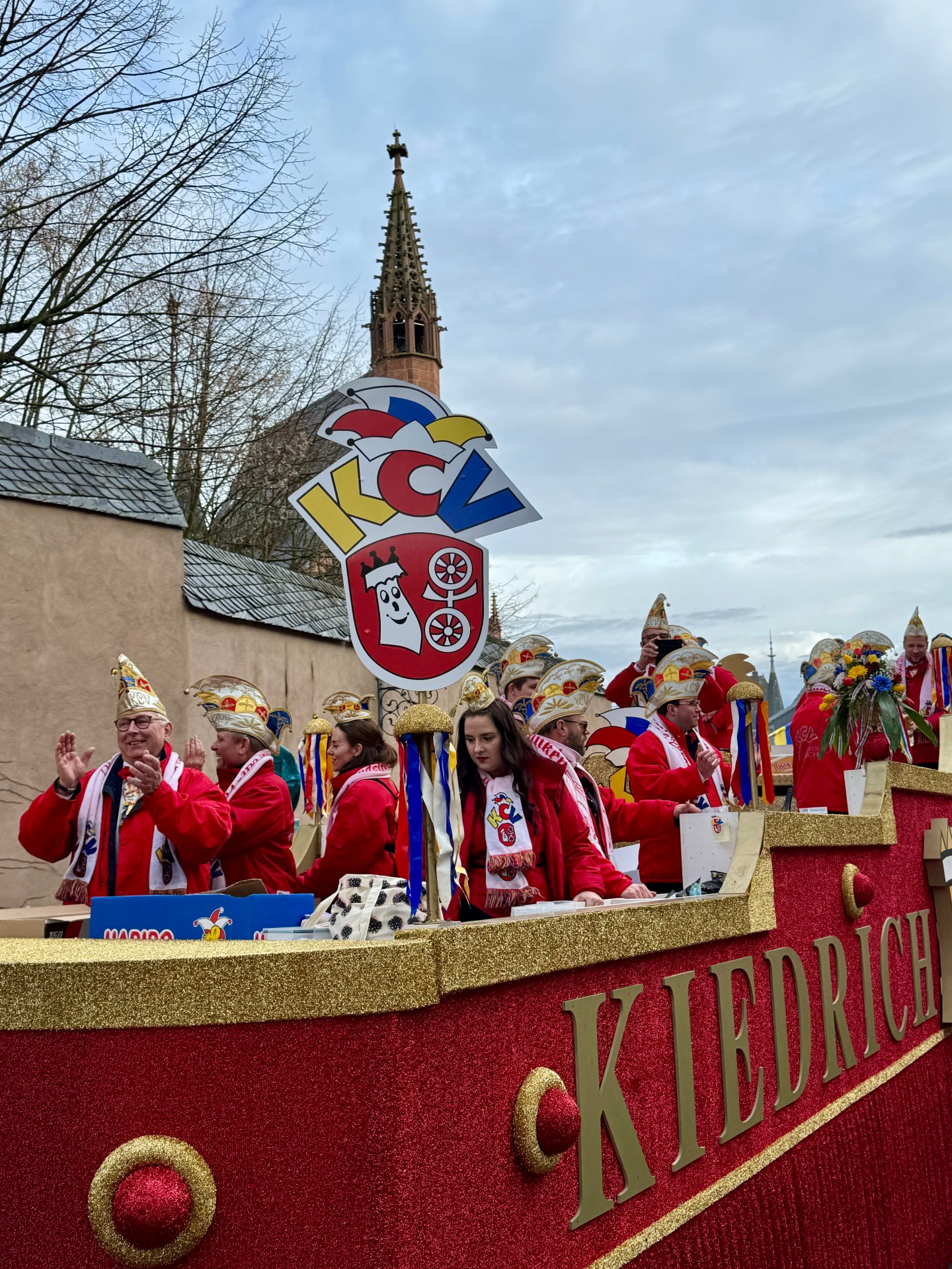 Ein Fastnachtswagen, der mit rotem und goldenem Glitzerstoff umhüllt ist. In Gold sind große Buchstaben, die das Wort Kiedrich bilden. Auf dem Wagen stehen mehrere Personen mit roten Jacken, weißen Schals mit dem KCV Logo und Narrenkappen auf dem Kopf. Es ist auf dem Wagen das große KCV Wappen in gelb, rot, blau mit den Buchstaben und der Kappe sowie dem Kiedricher Wappen in rot und dem Scharfenstein und dem Doppelrad in weiß zu erkennen. Im HIntergrund ist ein bewölkter Himmelund eine hellbraune Mauer und ein Baum ohne Blätter sowie ein Turm einer Kapelle in Sandsteinfarben. 