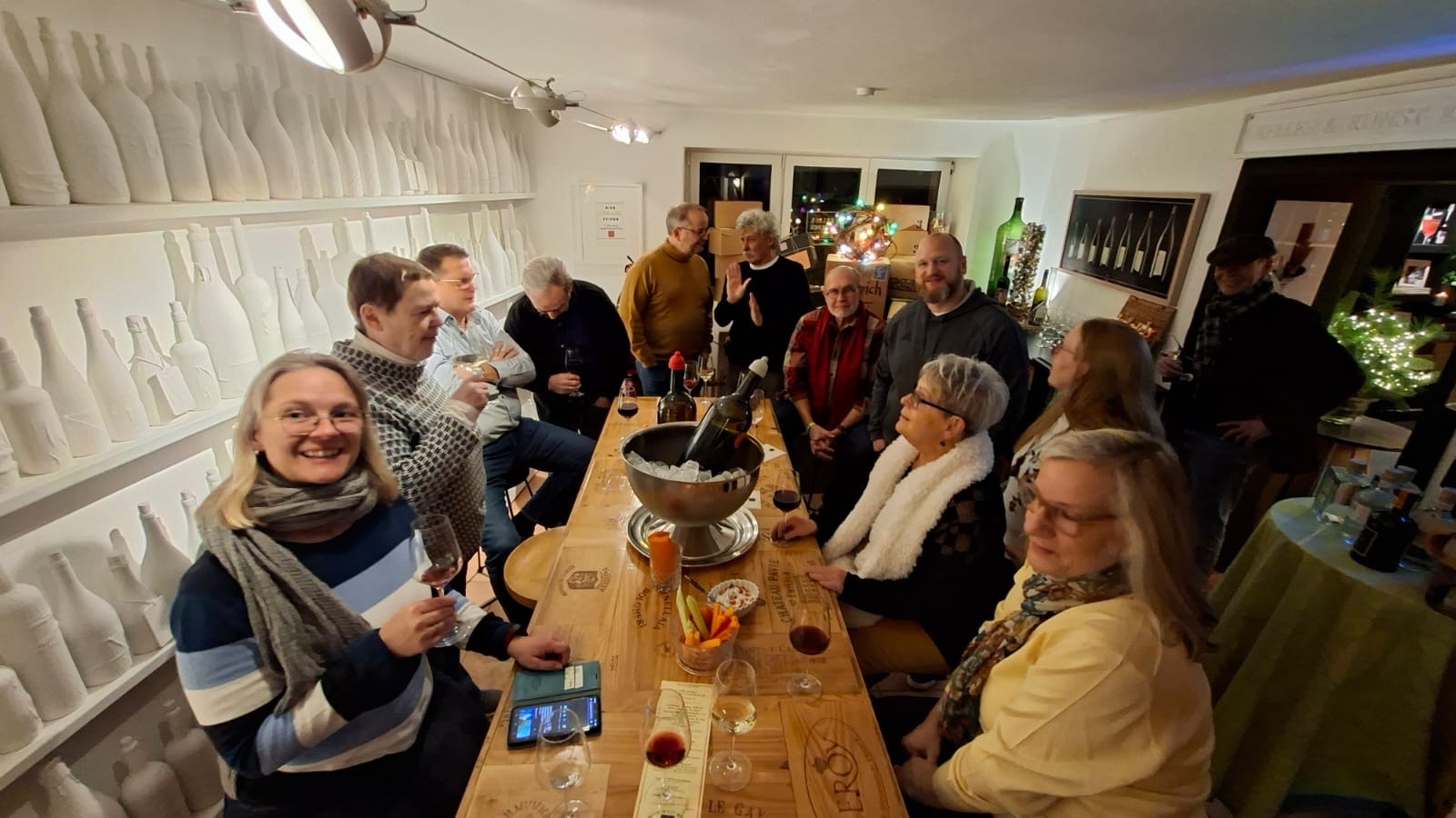 Links ist eine weiße Wand mit weißen Regalbrettern, auf denen weiße Flaschen dekoriert wurden. Man sieht einen langen Holztisch, auf dem gefüllte Weingläser und eine silberne Weinkühlerschale stehen. Um den Tisch sitzen und stehen Männer und Frauen.