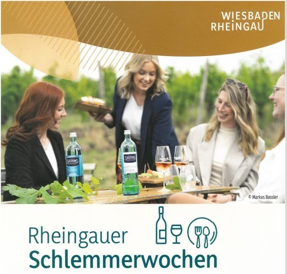 Plakatwerbung für Rheingauer Schlemmerwochen- Drei Frauen im Blazer sitzen an einem Tisch in den Weinbergen und eine Frau im Blazer reicht zwei Brettchen mit Essen an. Auf dem Tisch stehen Wasserglasflaschen und drei mit Rose gefüllte Weingläser und ein Wasserglas sowie Blätter eines Weinstocks.