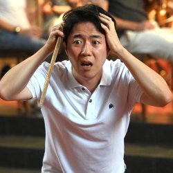 Ein Mann, mit asiatischen Gesichtszügen, hat ein weiße Poloshirt an und schlägt die Hände über den Kopf zusammen. In der einen Hand hält er einen Holzstab. Die Augen und der Mund sind aufgerissen.