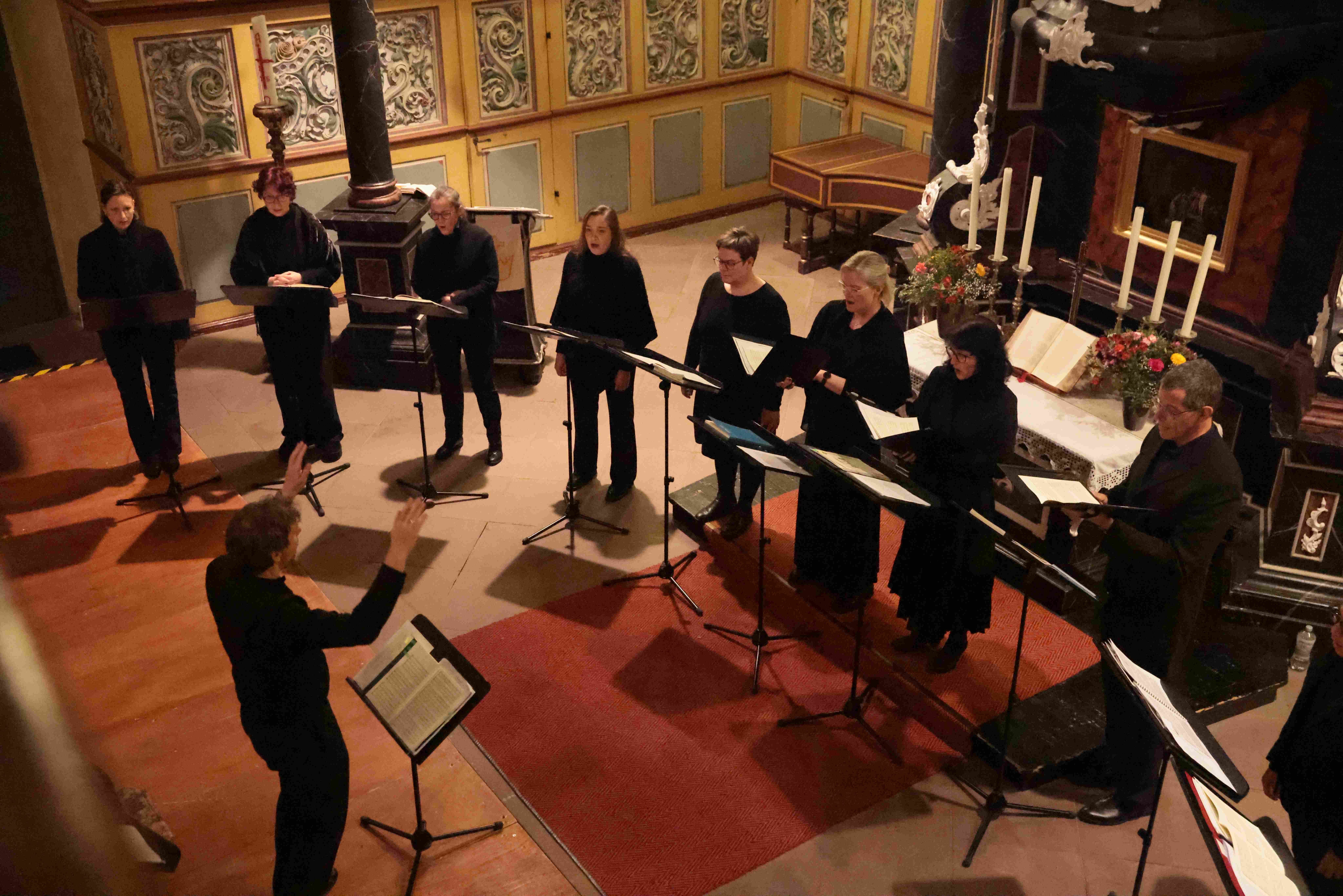 Blick von oben auf neun Personen, in schwarz gekleidet, die im Halbkreis hinter Notenständern stehen und singen. Davor steht ein Dirigent, ebenfalls in schwarz, der die Hände in die Höhe hält und ebenso einen Notenständer vor sich hat. Die Personen stehen in einem Altarraum mit Blumenschmuck und Kerzen und verzierten Wänden. 