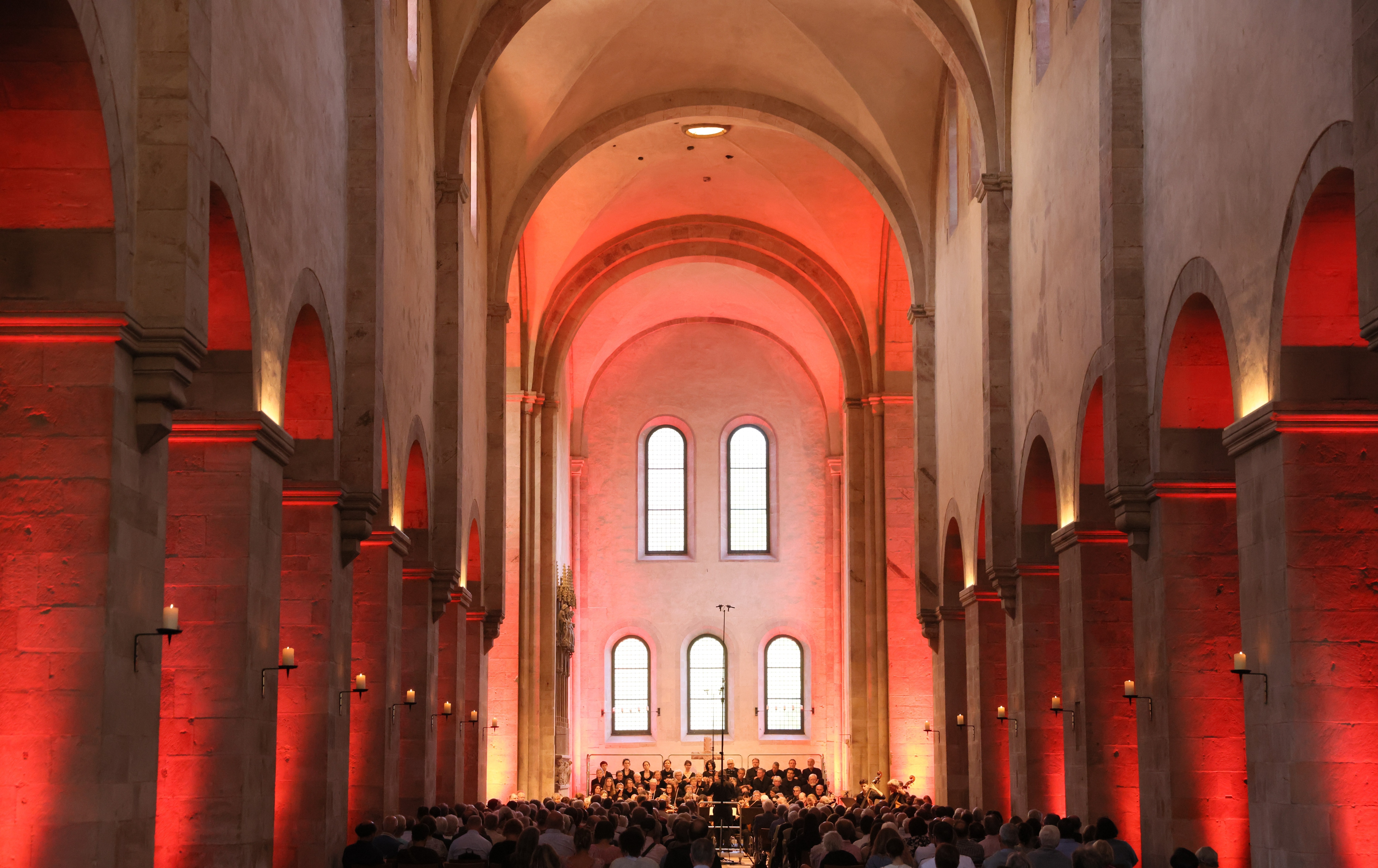 Blick auf ein Kirchenschiff. Seitlich sind hohe Säulen mit Rundbögen. An den Säulen sind beige brennende Kerzen auf schwarzen Kerzenhaltern. Die Säulen sind rot angestrahlt. Es sitzt dort ein Publikum, welches auf einen schwarz gekleideten Chor blickt. Dahinter sind drei Fenster mit Rundbogen oben und darüber zwei größere Fenster mit Rundbogen.  