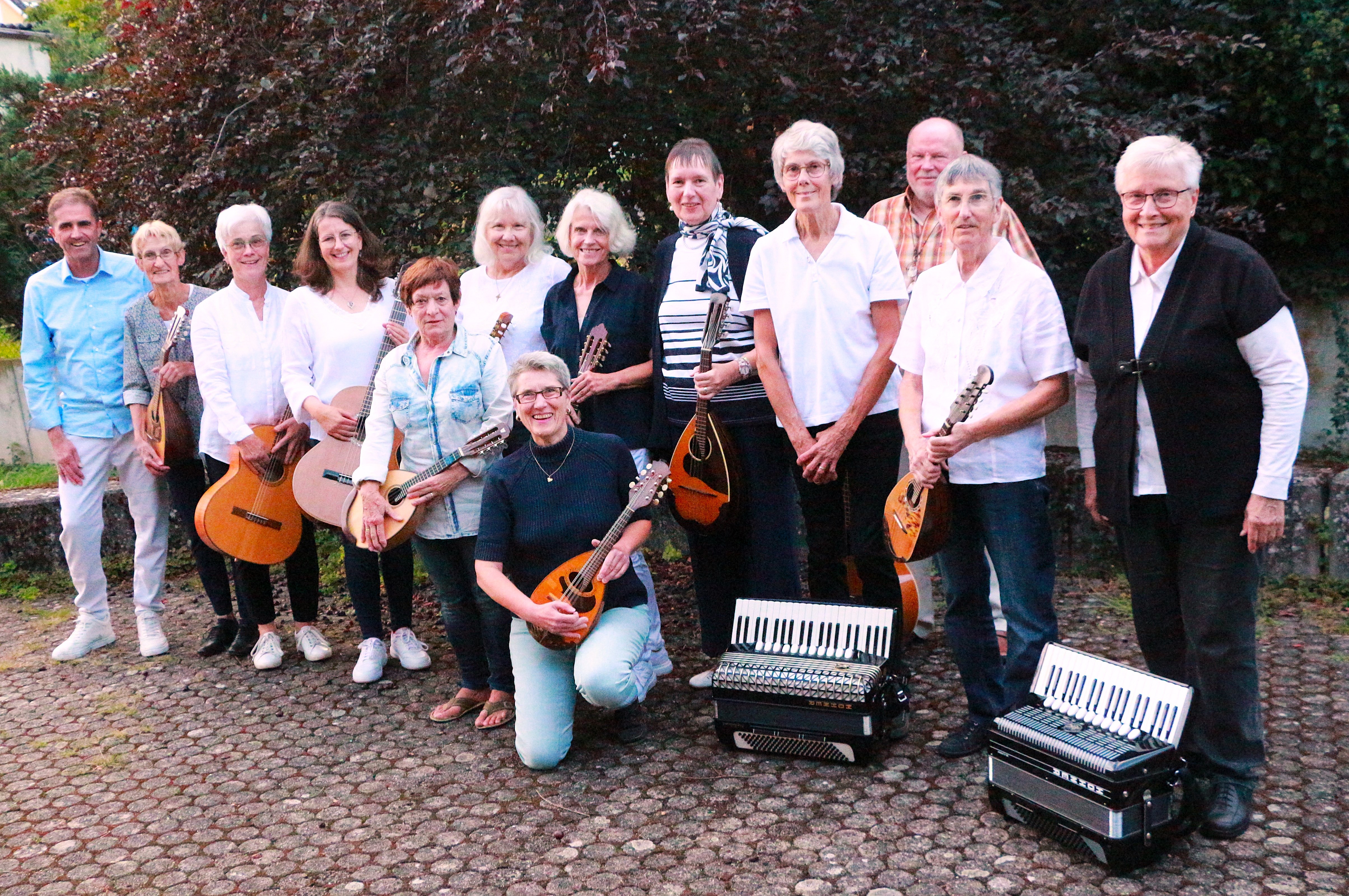 Auf einer Fläche, die mit fünfeckigen Steinen gepflastert ist, stehen vor einem Laubbaum mit roten Blättern, 13 Personen, zum Teil mit Mandolinen und einer Gitarre in den Händen. Davor liegen zwei Akkordions.  