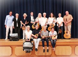 Auf einer Bühne mit braunem Holzboden und brauner Verkleidung, stehen vor einem blauen Bühnenvorhang 9 Personen und davor sitzen am Bühnenrand 4 Personen, die die Beine baumeln lassen. Die Personen haben Mandolinen, Gitarren und Akkordions in den Händen.  