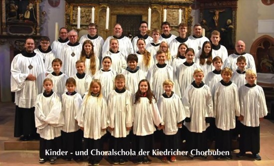 Männer der Kiedricher Chorbuben und der Kinder- und Chorschola stehen in ihren weiß- schwarzen Chorröcken vor dem Altar auf dem sechs lange, weiße Kerzen stehen.