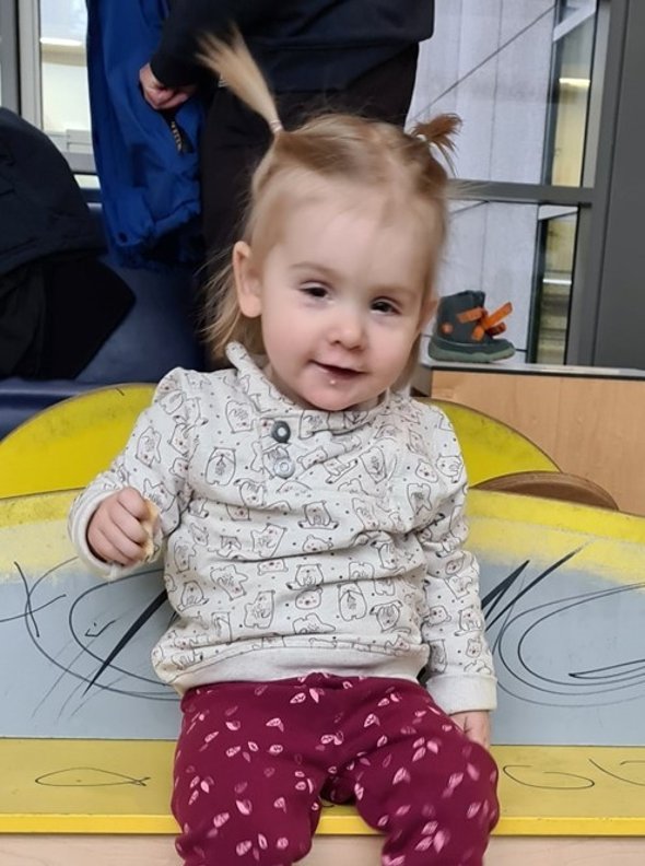 Marleen Ein kleines blondes Mädchen mit zwei Zöpfen mit pinker Hose und beigem Pullover, jeweils mit Muster, lacht in die Kamera.