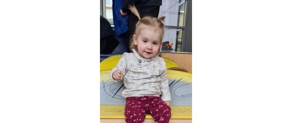 Ein kleines blondes Mädchen mit zwei Zöpfen mit pinker Hose und beigem Pullover, jeweils mit Muster, lacht in die Kamera.
