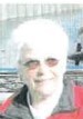 Eine Frau mit weißen, kurzen Haaren, mit einer Sonnenbrille, und einer roten Jacke mit schwarzem Kragen vor einem hell- und dunkelgrauem und blauem Hintergrund.