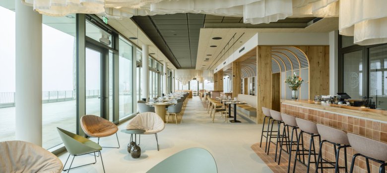 Ein langer, großer Raum, links und hinten ist eine Gläserfront. Rechts vorn ist eine Bar mit viereckogen Holzverkleidung. Die BArhocke haben schwarze Metallbeine und beige Sitzschalen. Links stehen Loungesessel in beige, braun und grün. Im HIntergrund lange Tische und Sitzbänke sowie Stühle. Die Decke ist unterschiedlich, u.a. im Vordergrund Stoff, der weiße Wellen darstellt.