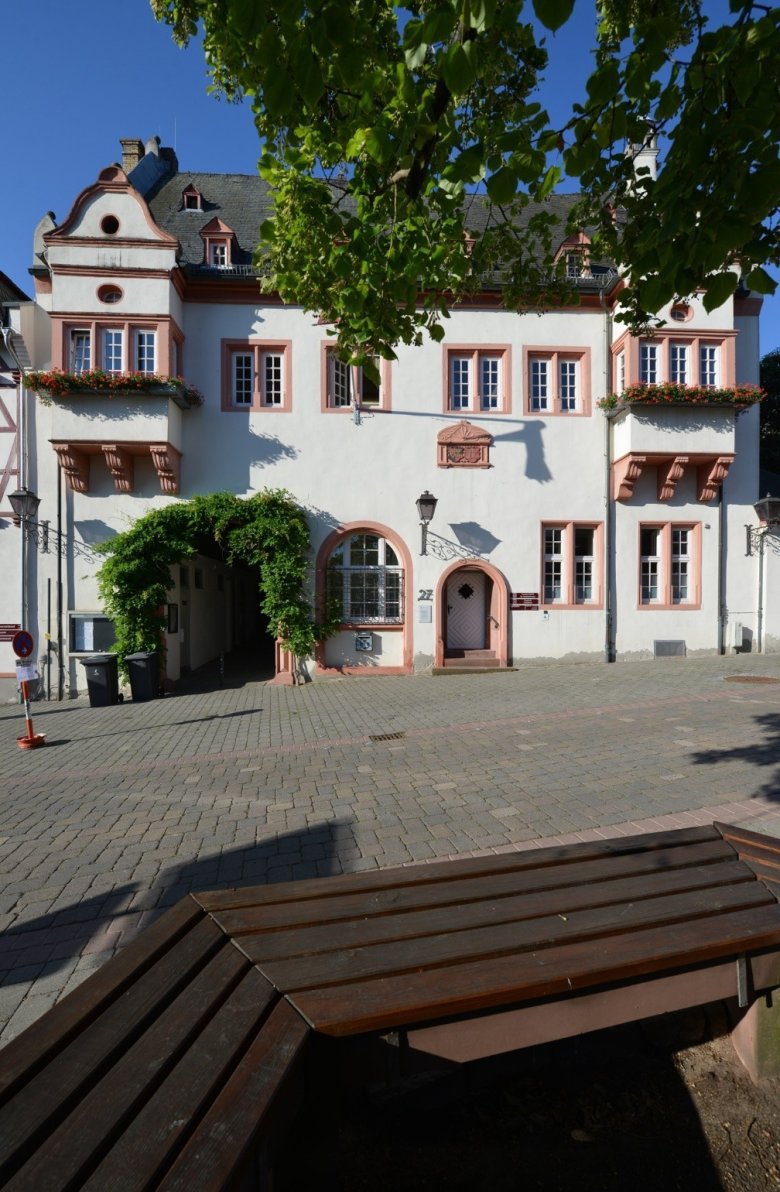 Kiedricher Rathaus, weisses Gebäude mit Sandsteinfensterrahmen, zwei Erkern, dem Eingang und drei Stufen aus Sandstein. Links von der Eingangstür, neben einem großem oben rundem Sprossenfenster, ist ein mit grünen Blättern umrankter Torbogen. Oben im Bild sieht man ein paar Blätter an Ästen und unten eine Holzbank mit Ecken.