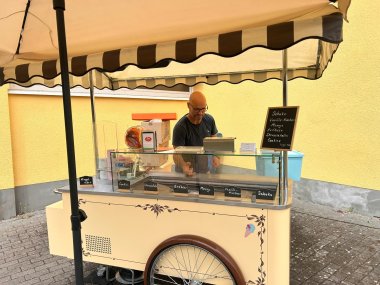 Ein beiger Eiswagen mit einem braunem Speichemrad und einem beige-schwarz gestreiftem Schirm, dahinter ein Mann und eine Tafel mit den Eissorten. Der Hintergrund ist eine gelbe Mauer.