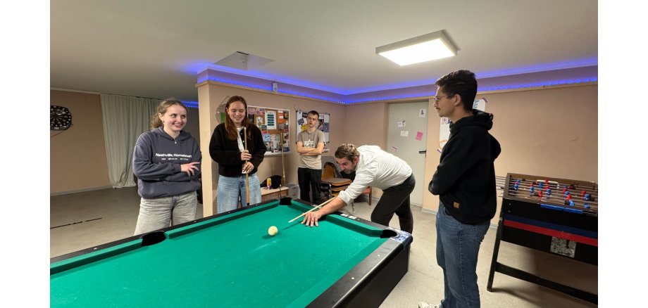 In einem großem Raum hängt eine Dartscheibe und eine Pinwand an der Wand und es steht ein Bililardtisch und ein Tischkicker im Raum. Um den Tisch stehen vier Jugendliche und ein Mann, der den Kö in der Hand hält und kurz davor ist, mit dem Stoß zur weißen Kugel, das Spiel zu beginnen. 