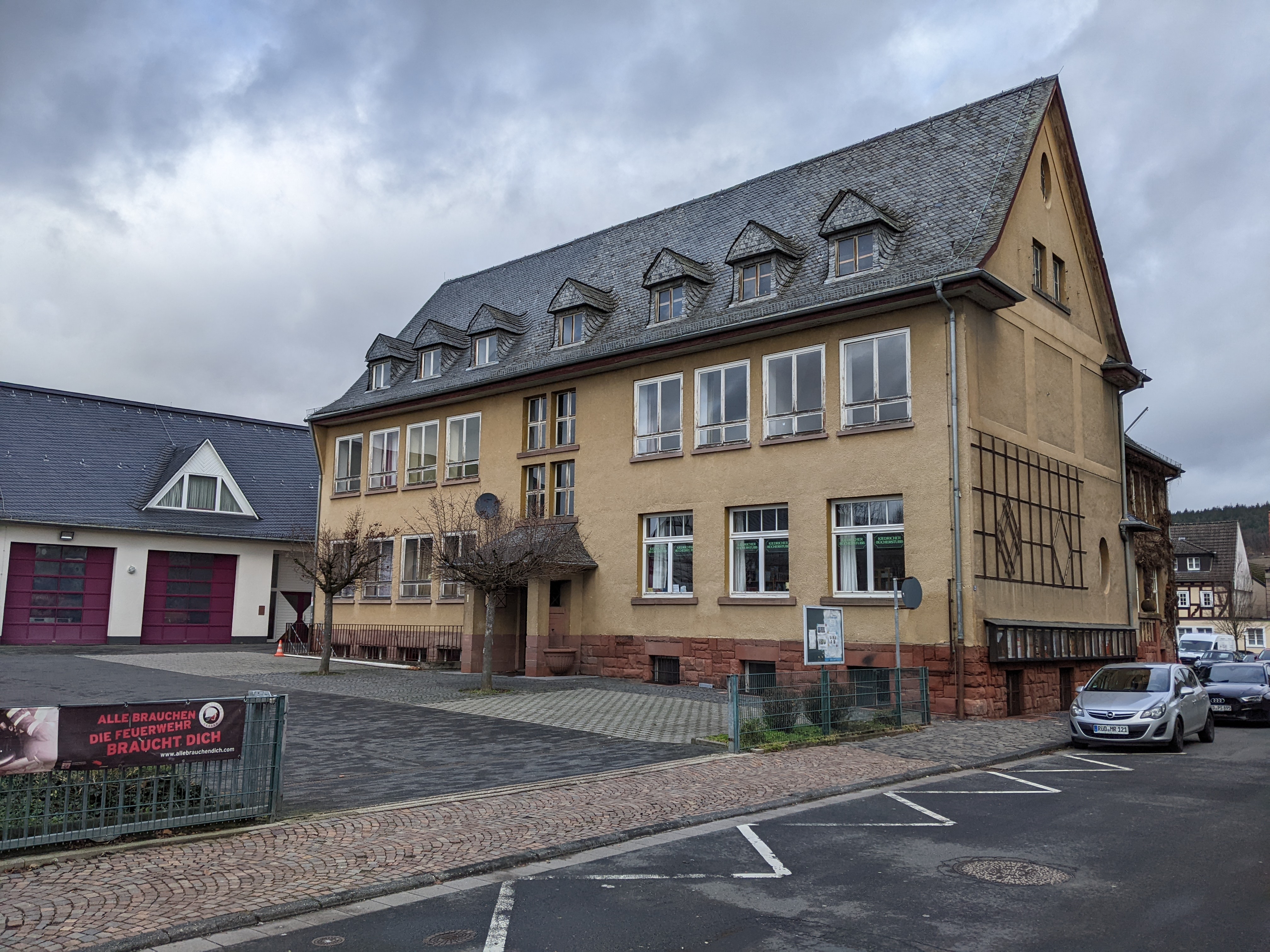 Beiges Gebäude mit Fenstern und Dachfenstern in dem mit schwarzen Ziegeln bedeckten Dach, In dem Gebäude befand sich früher die Schule von Kiedrich. Ebenso sieht man den Hof mit Bäumen und ein Teil der Straße mit einem parkendem Auto. 