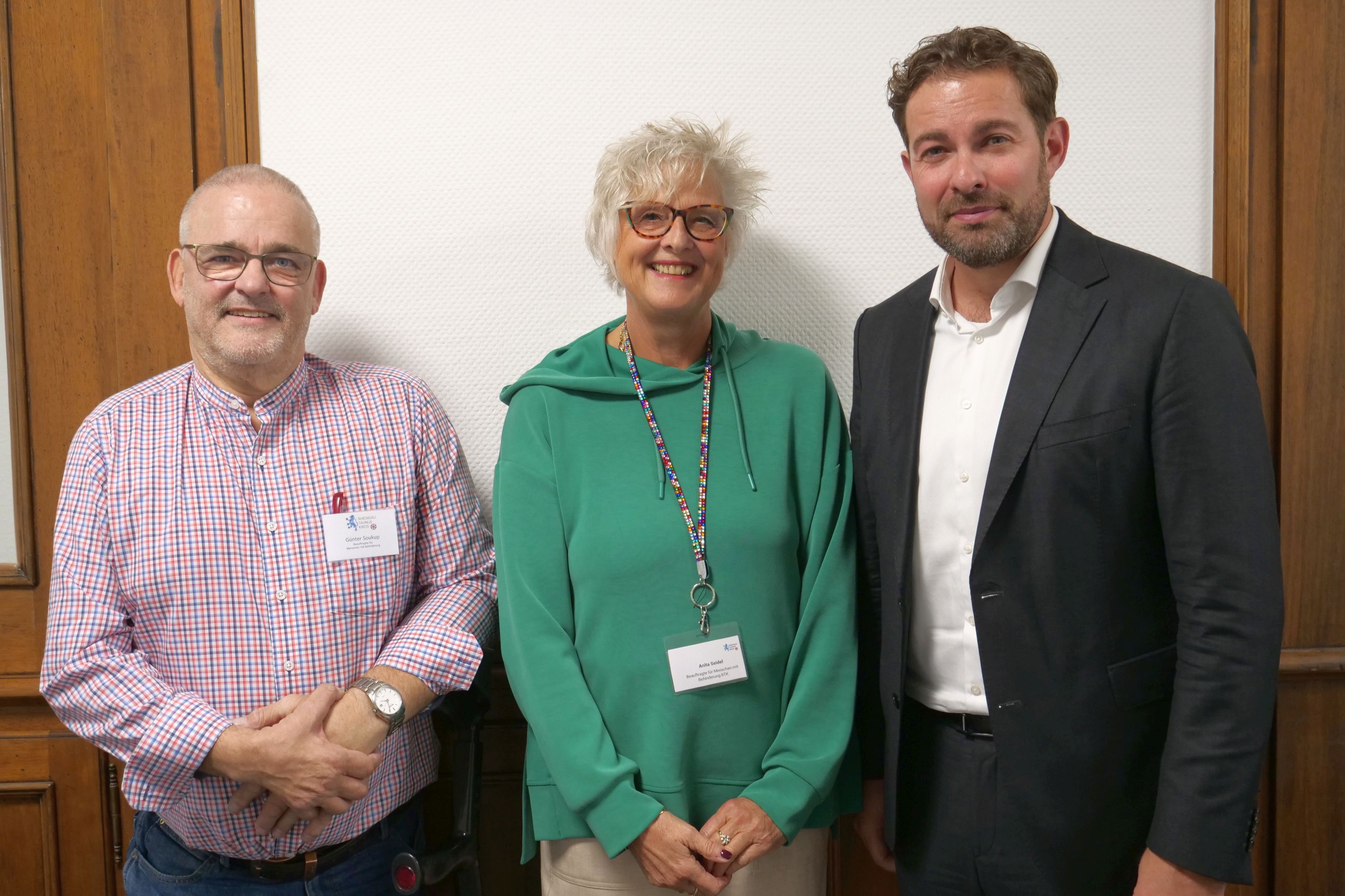 Eine Dame mit blonden Locken und grünem Pullover steht in der Mitte von einem Mann mit Anzug und weißem Hemd und einem Mann mit einem rot karriertem Hemd.
