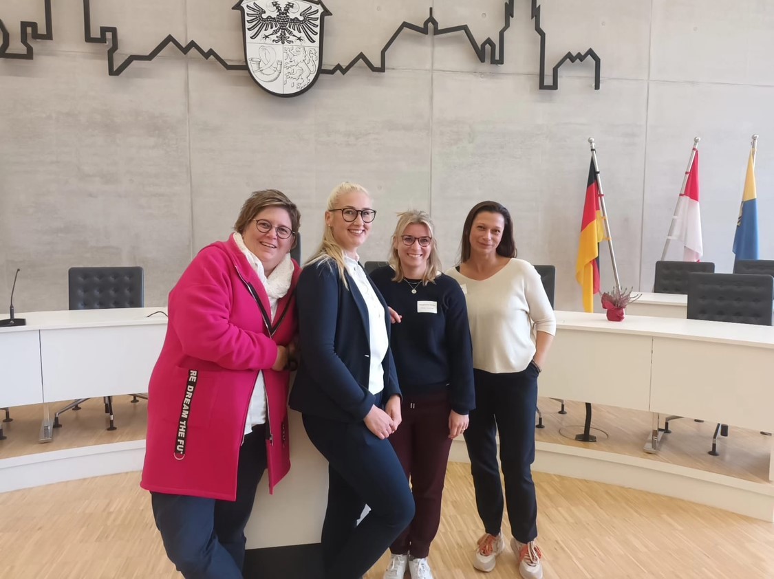 Zwei braunhaarige und zwei blonde Frauen, drei davon tragen eine Brille, stehen vor einem Halbkreis mit weißen Tischen mit Blende, einem Mikrofon und einer rosa Topfplume auf den Tischen. Die Schreibtischstühle sind schwarz. Am rechten Bildrand sind drei Fahnen und am oberen Bildrand die Siluette einer Stadt und ein schwarz-weißes Wappen.