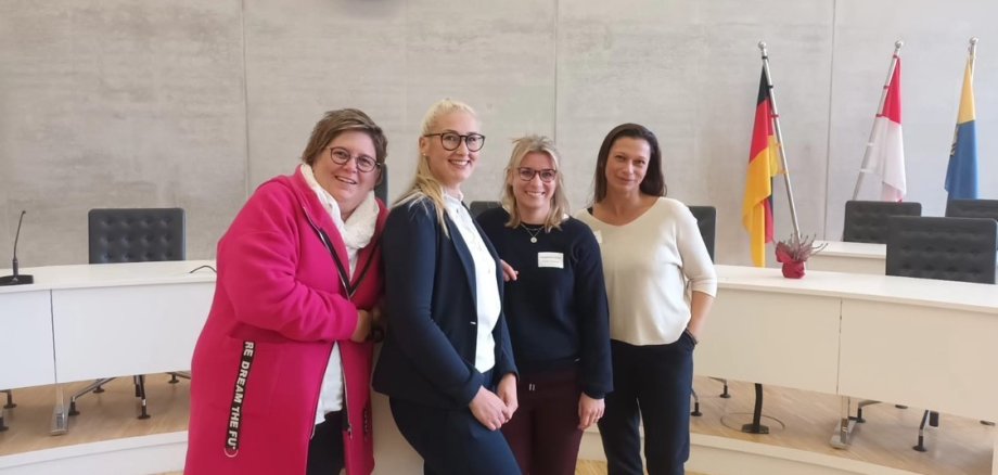 Zwei braunhaarige und zwei blonde Frauen, drei davon tragen eine Brille, stehen vor einem Halbkreis mit weißen Tischen mit Blende, einem Mikrofon und einer rosa Topfplume auf den Tischen. Die Schreibtischstühle sind schwarz. Am rechten Bildrand sind drei Fahnen und am oberen Bildrand die Siluette einer Stadt und ein schwarz-weißes Wappen.