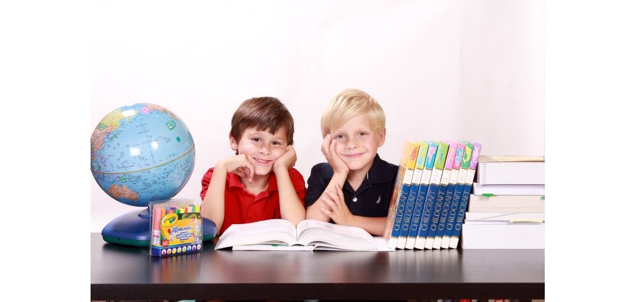 Ein blonder und ein braunhaariger Junge sitzen an einem Tisch und stützen ihre Köpfe auf den Händen ab. Vor ihnen liegt ein aufgeschlagenes Buch und auf dem Tisch steht ein Globus, weitere Bücher und Stifte.