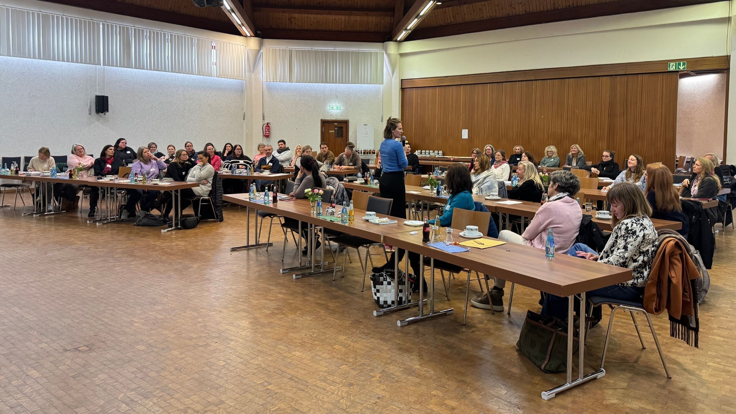 In einem Saal mit einem Holzboden sind jeweils 2x drei Tische mit Lücke nebeneinander gestellt in vier Reihen. An den Tischen sitzen mehrere Personen.