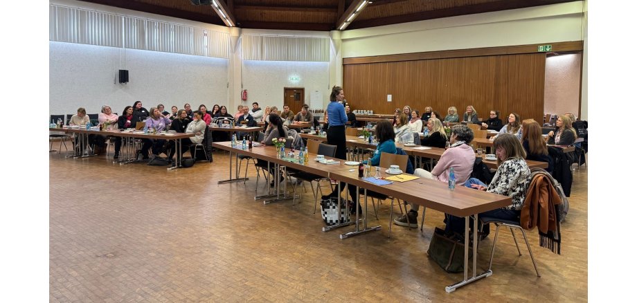 In einem Saal mit einem Holzboden sind jeweils 2x drei Tische mit Lücke nebeneinander gestellt in vier Reihen. An den Tischen sitzen mehrere Personen.