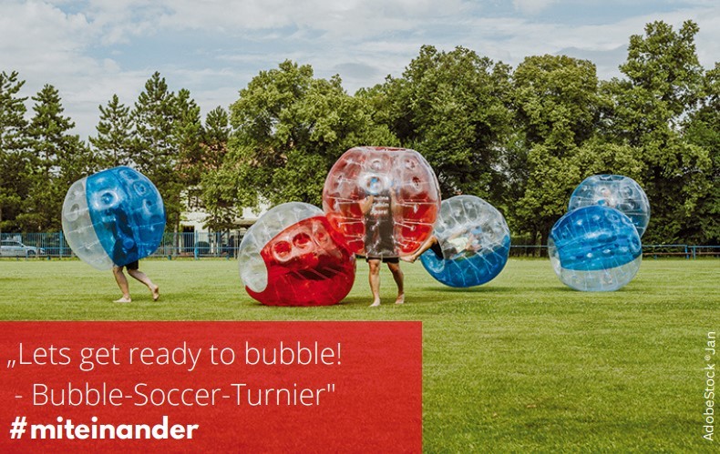 Auf einer Rasenfläche, mit Bäumen im Hintergrund sind zwei Menschen mit Kopf und Oberkörper in einer rot und durchsichtigen Bubble und einer in einer blau und durchsichtigen Bubble. Auf dem Rasen liegt eine weitere rote und drei blaue Bubble-Bälle.