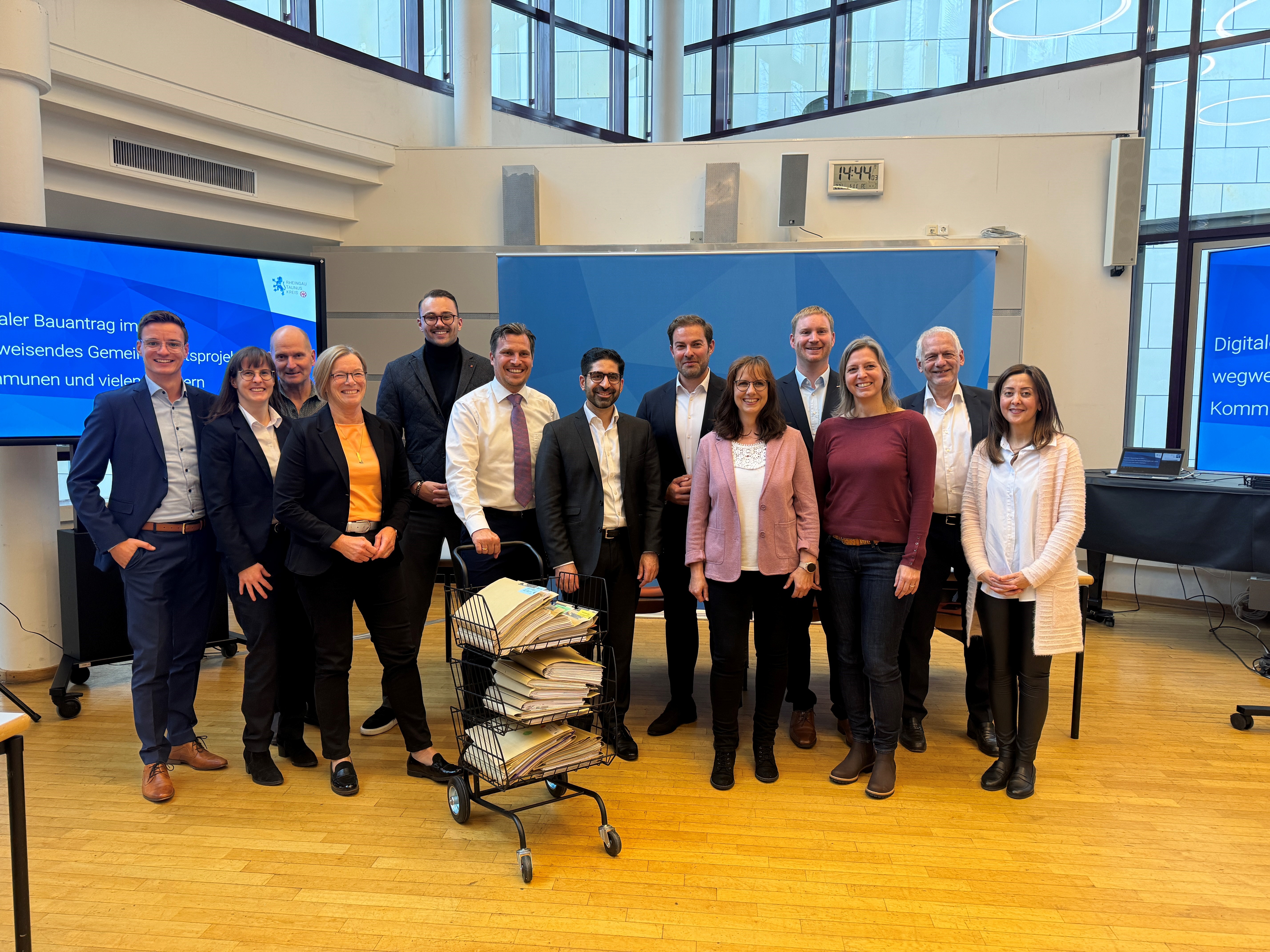 Fünf Frauen und acht Männer stehen im Kreishaus vor zwei  großen Bildschirmen und einer Leinwand hinter einem dreikörbigen, schwarzen Aktenwagen, der mit braunen Aktenordnern gefüllt ist. 