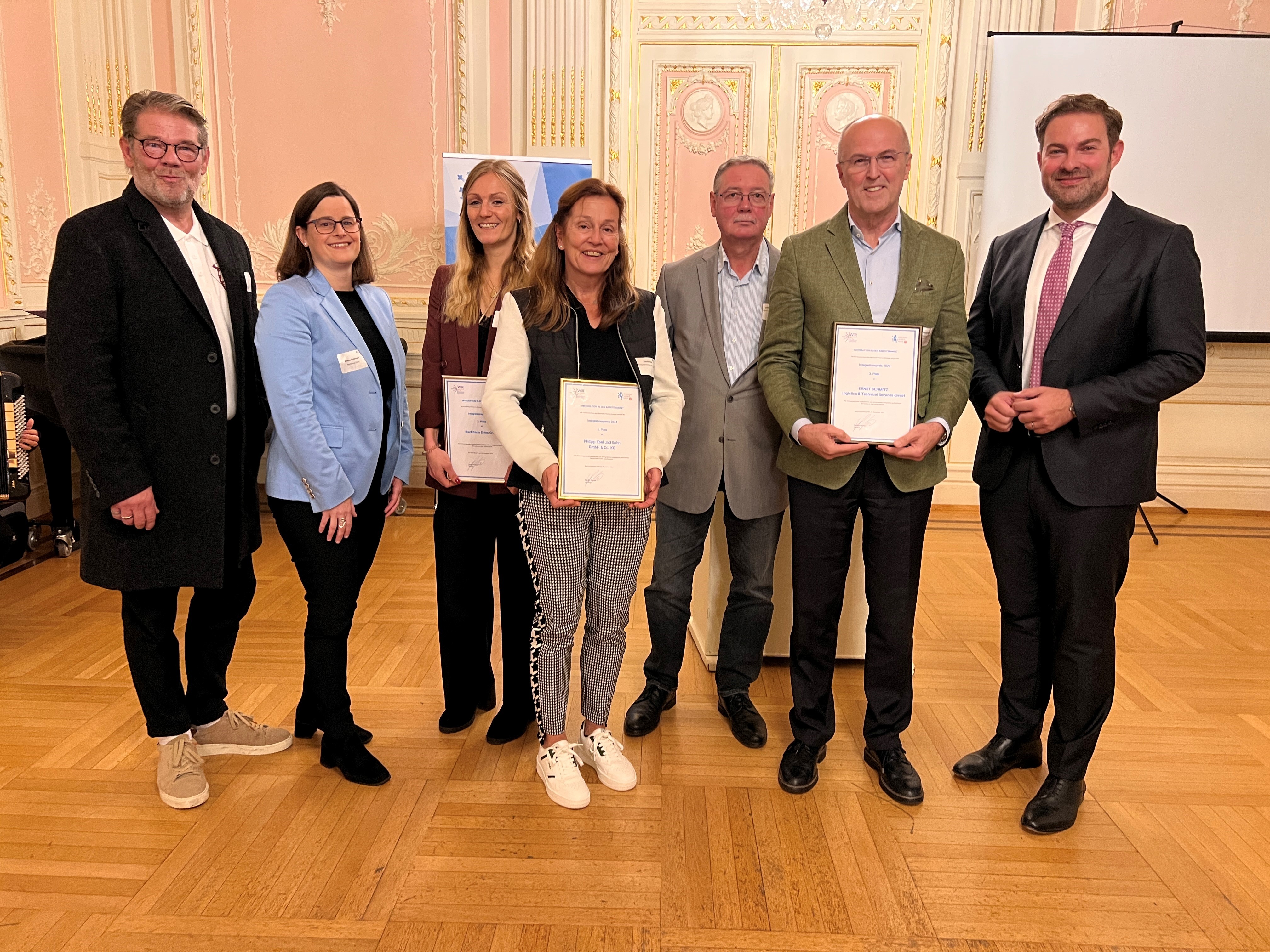 Vier Herren und drei Damen stehen nebeneinander. Drei davon halten eine Urkunde in der Hand. Im Hintergrund eine rosa Wand und Tür, die mit weißem Stuck und goldenen Verzierungen ausgestattet ist.