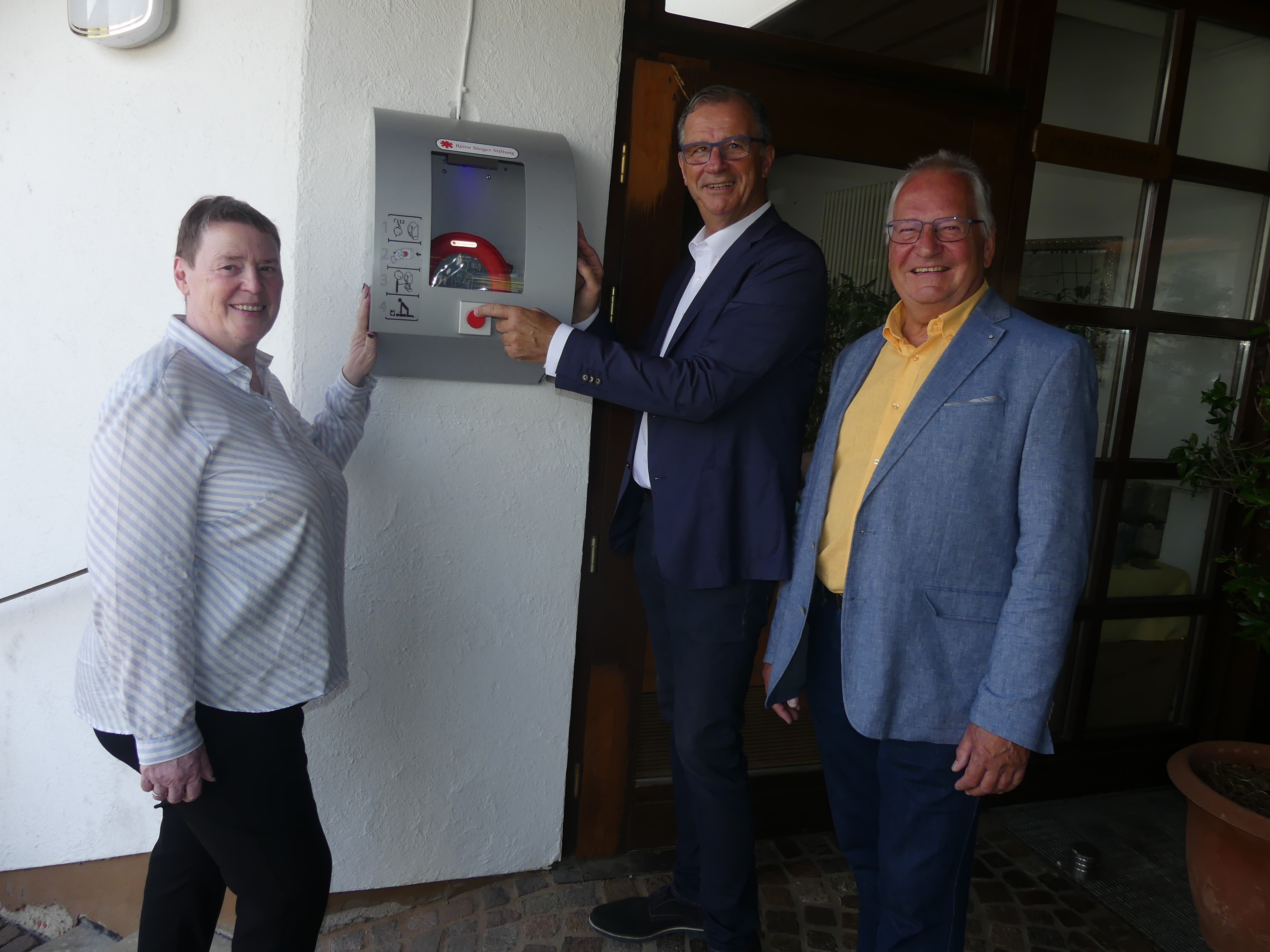 An einer weißen Wand hängt ein silbernen Kasten indem ein Defibrilator angebracht ist. Links davon steht Beate Schmidt in dunkler Hose und weißer Bluse und rechts davon Bürgermeister Steinmacher im dunklem Anzug und weißem Hemd und Rüdiger Wolf im gelben Hemd, hellblauem Sakko und dunkler Hose.