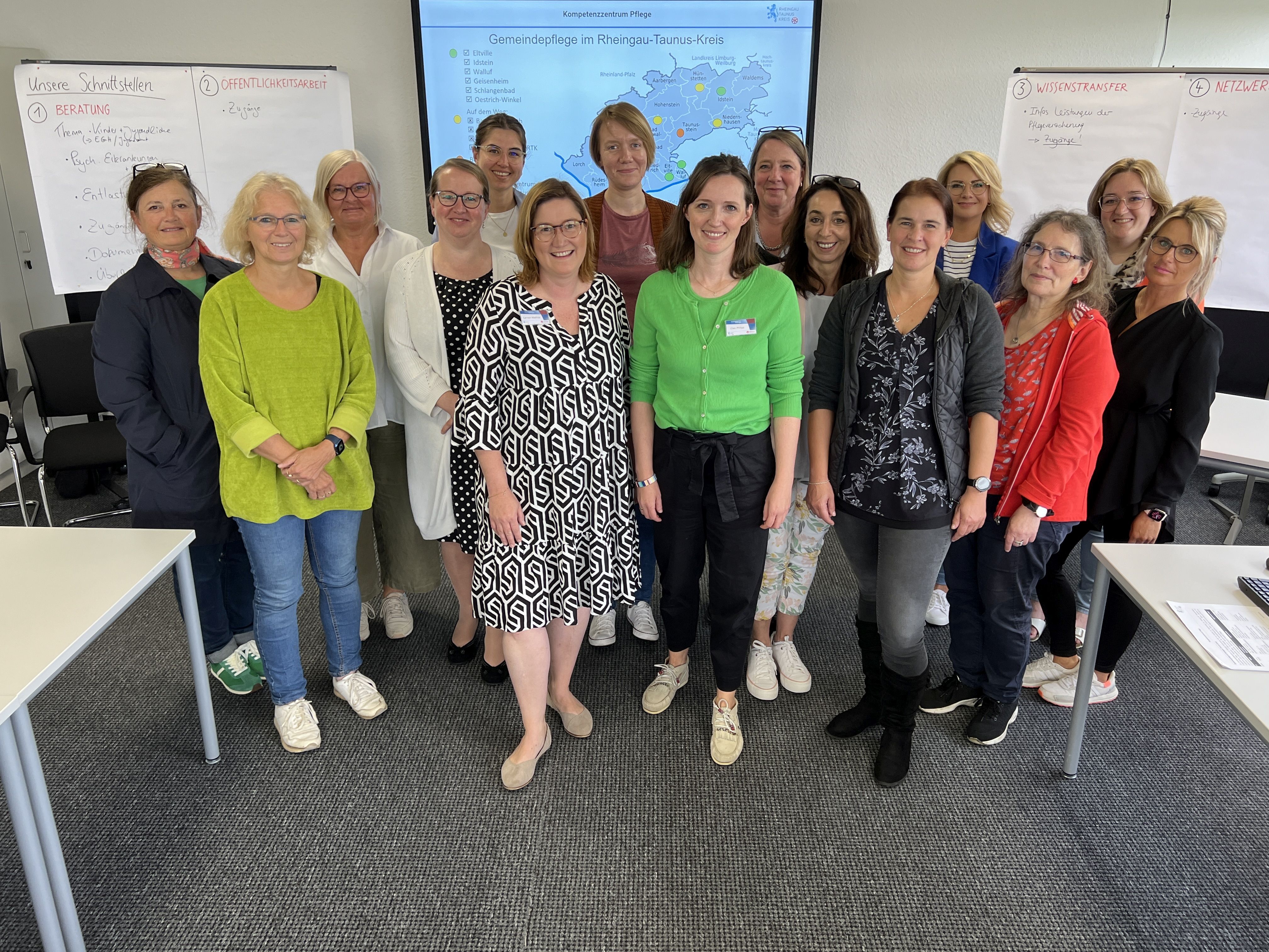 Vor einer Leinwand mit einer Karte, neben der links und rechts mehrere Flip-Charts mit beschrifteten Blättern hängen, stehen  15 Frauen.