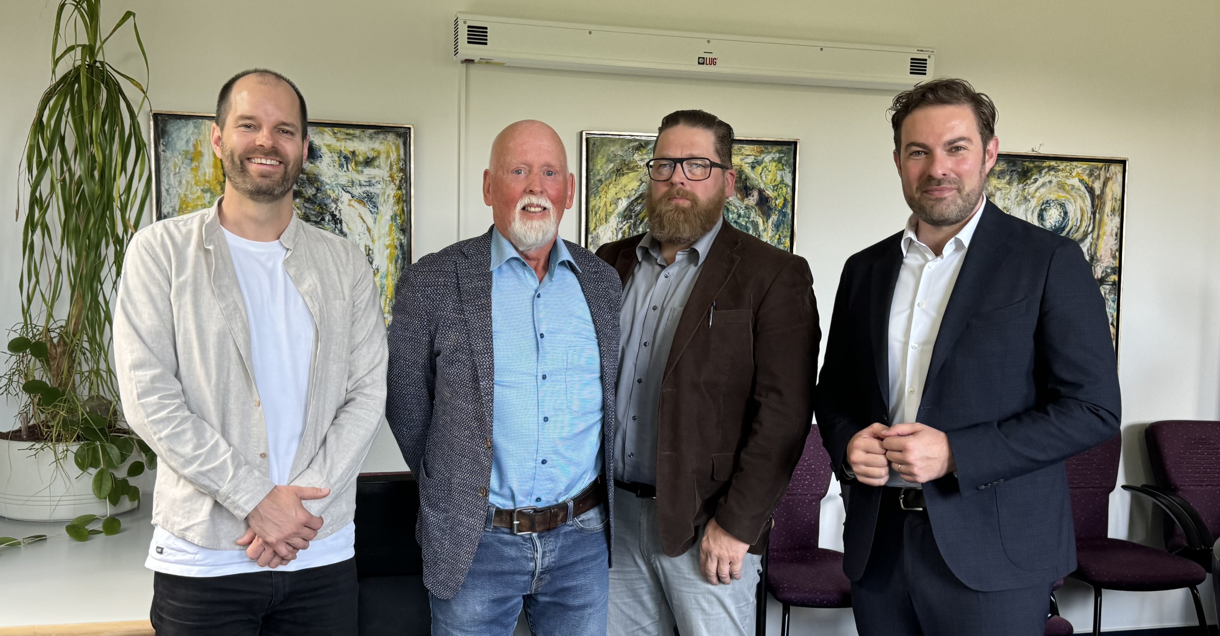 Vier Männer stehen nebeneinander vor einer Topfblume und drei Gemälden, die an der Wand hängen.