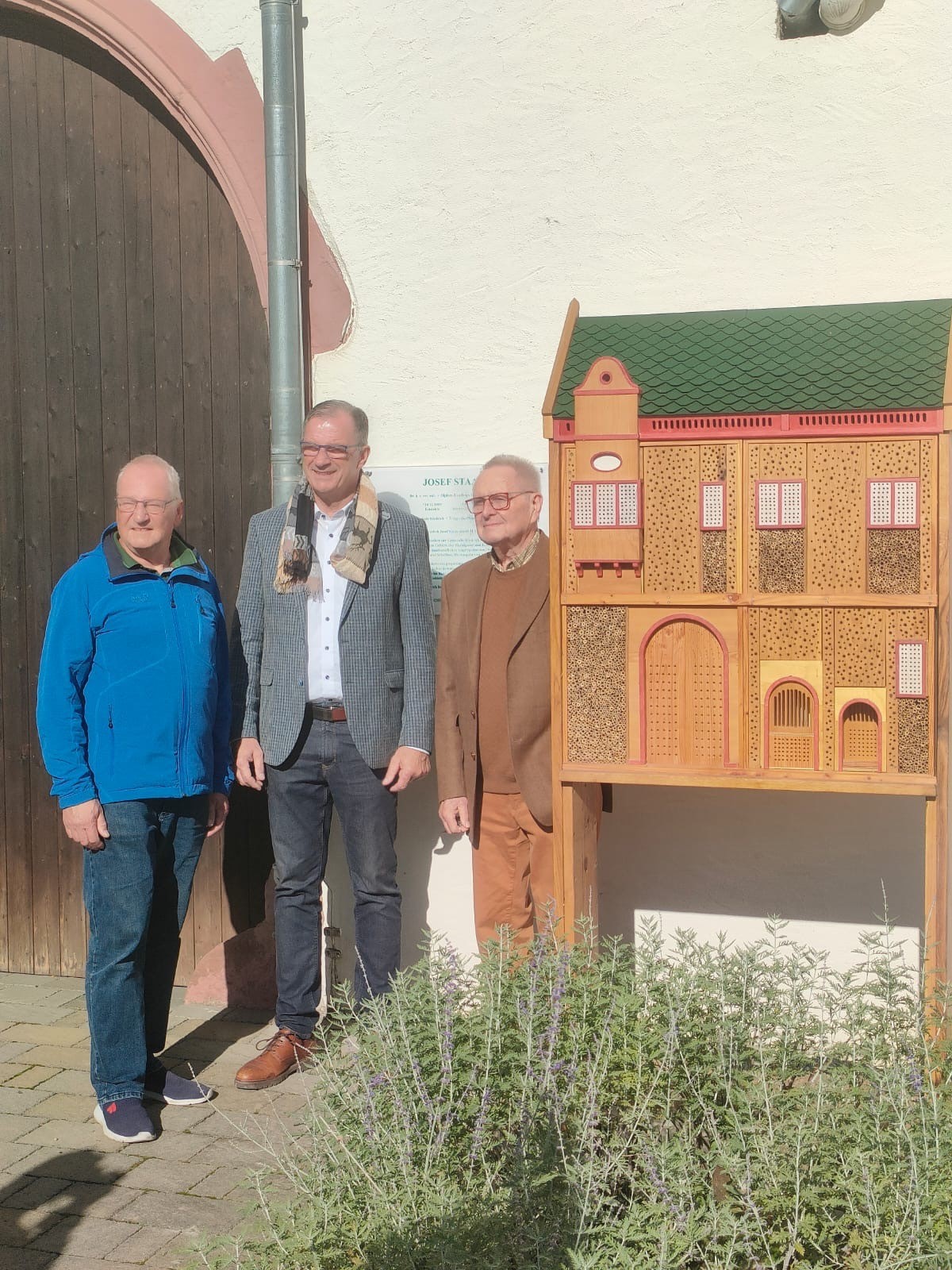 Erster Beigeordneter Rüdiger Wolf (links), Bürgermeister Winfried Steinmacher (Mitte), Dieter Steven (rechts) stehen neben dem neu aufgestelten Insektenhotel, welches mit Holz gebaut wurde und grüne Dachplatten und rote Fenster- und Türrahmen und verschiedenen Erkern besitzt und somit dem Kiedricher Rathaus ähnelt.