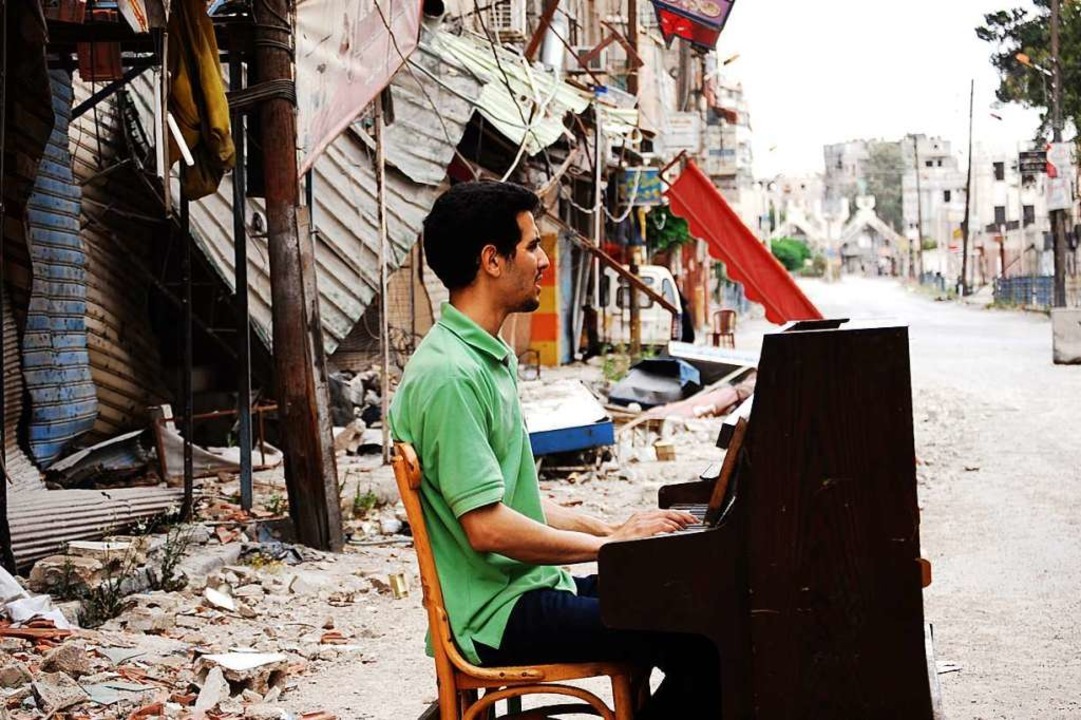 In einer zerbombtenStraße sitzt ein junger Mann mit dunklen Haaren und einem grünen Poloshirt und einer schwarzen Hose auf einem braunen Stuhl an einem schwarzen Klavier und spielt darauf. 