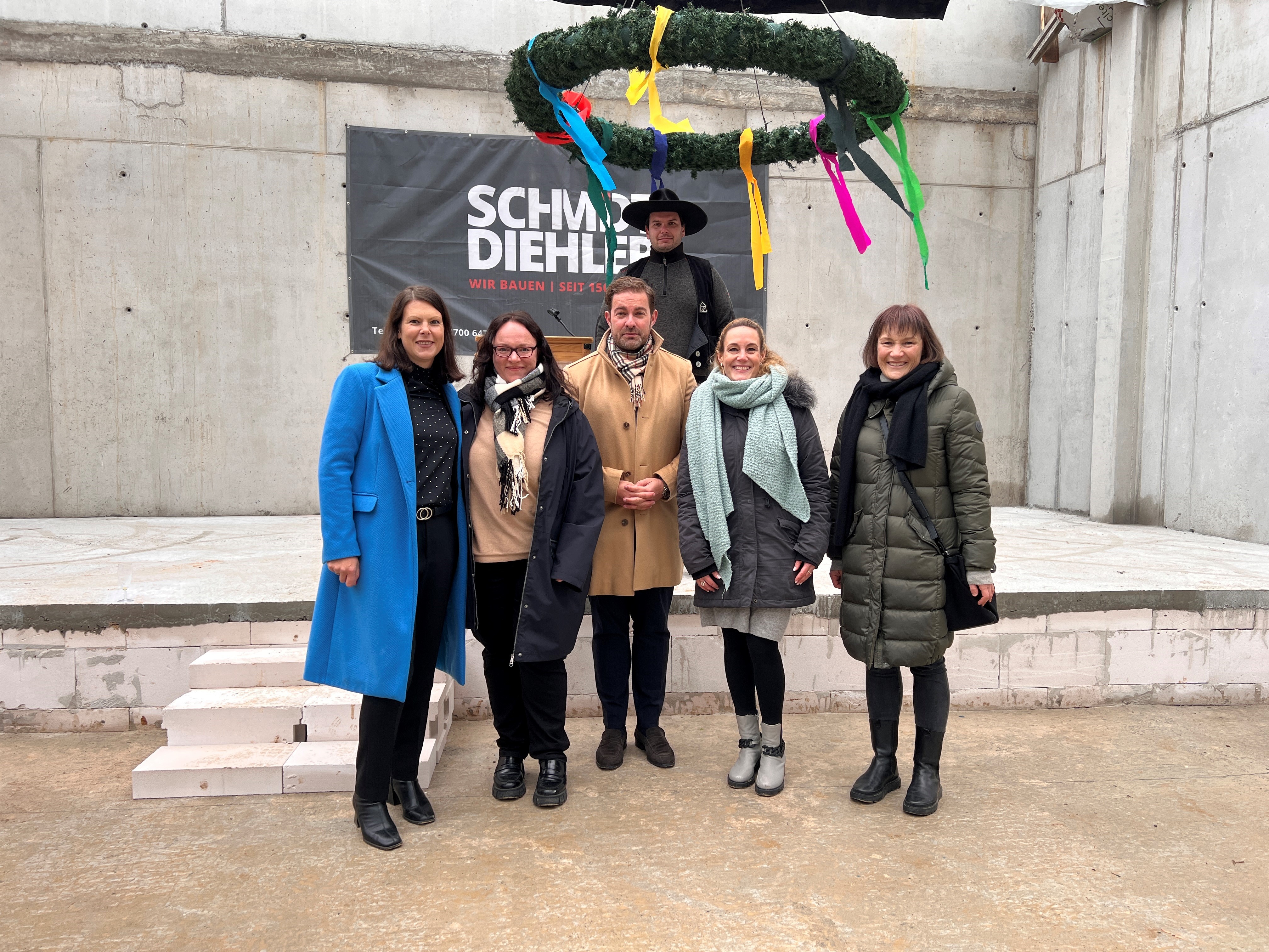 Vor einem Betonboden und Betonwänden und einer dreistufigen Treppe stehen unter einem Richtfestkranz mit bunten Bändern vier Damen und zwei Männer mit Winterjacken. 