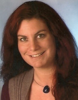 Frau mit langen braunen Haaren, einer Kette mit einem schwarzem Stein und einer braunen Strickweste mit Streifen. 