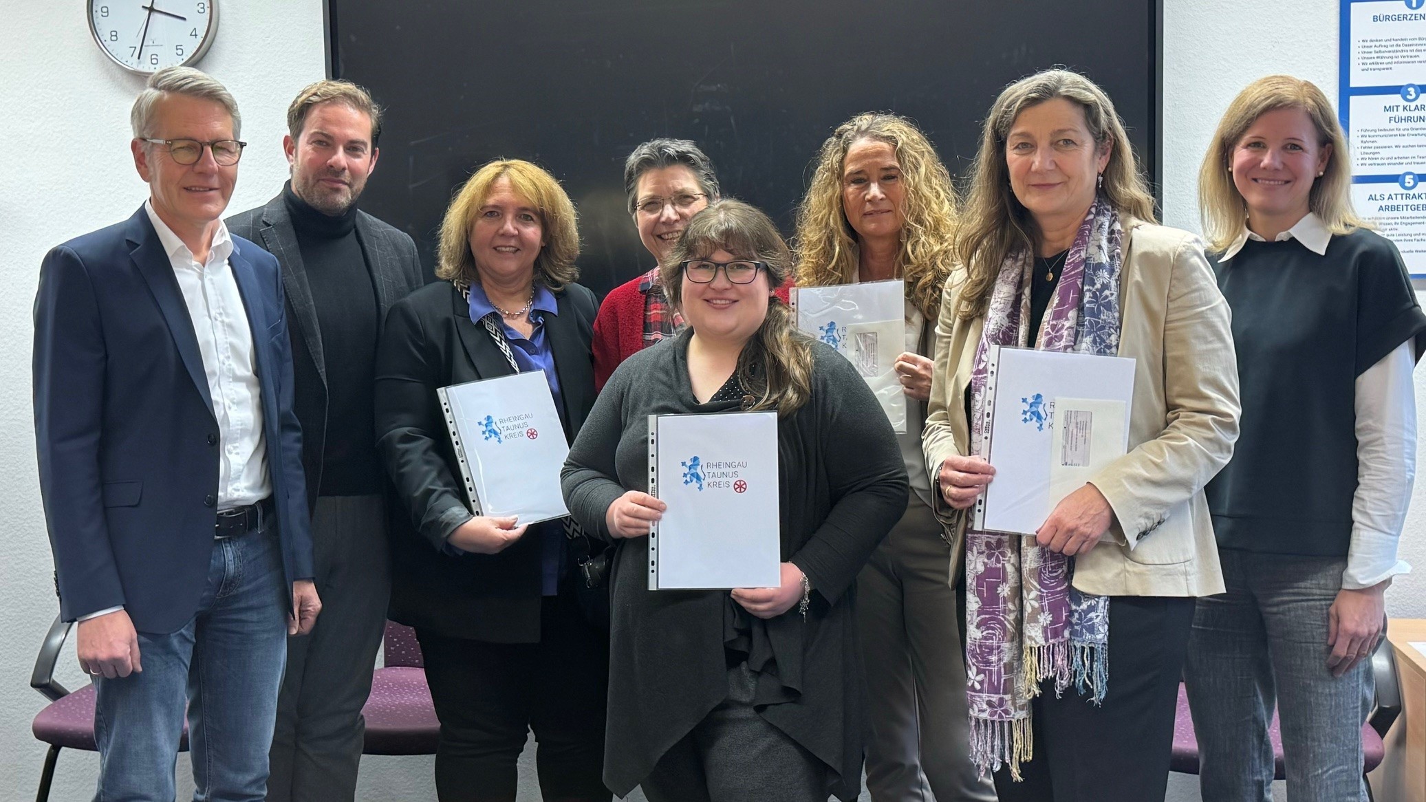 Vor einer Tafel und einer Wanduhr stehen Landrat Zehner mit einem Mann und sechs Frauen, die teilweise Urkunden des Rheingau-Taunus-Kreises in der Hand haben.