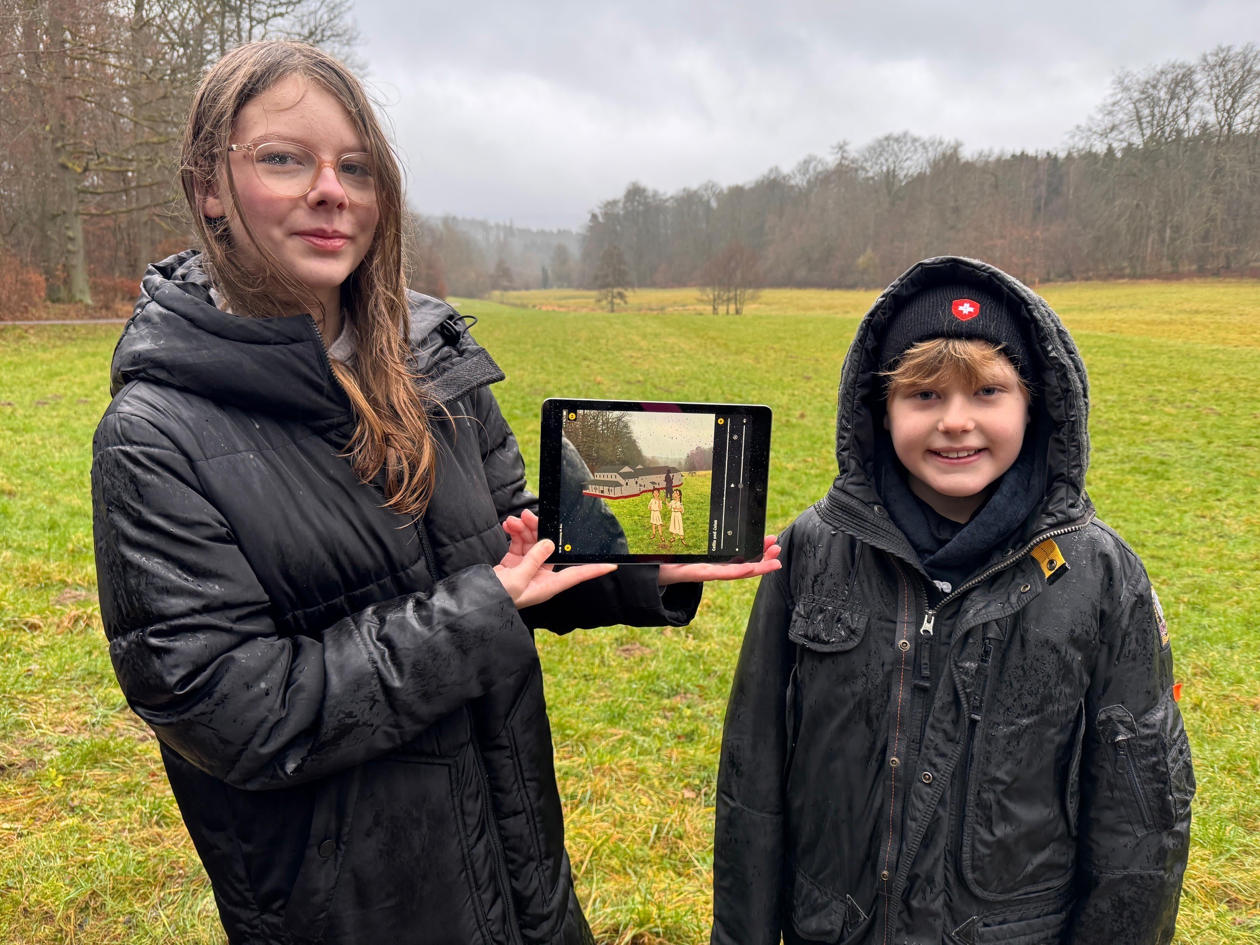 Ein Junge und ein Mädchen stehen mit schwarzen Winterjacken vor einer Wiese, im Hintergrund sind Bäume und halten ein Tablett, auf dem gezeichnet ein römisches Gebäude und zwei Kinder im Römer-Gewand zu sehen ist.