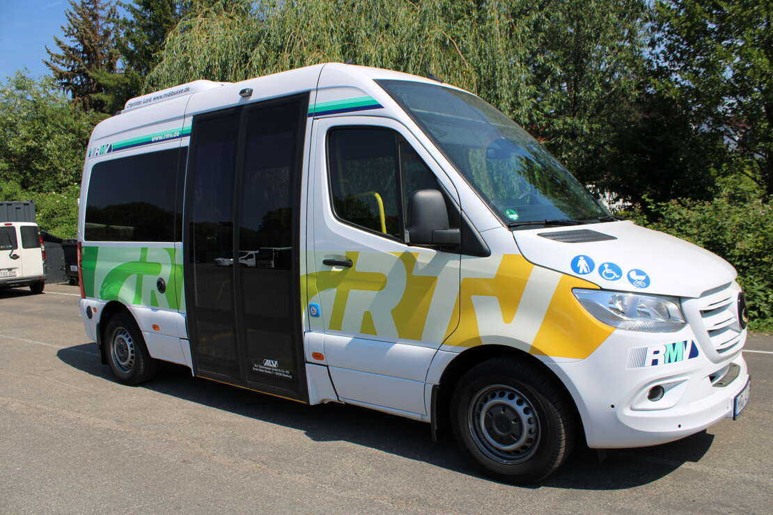 Ein weißer kleiner Bus mit dunklen Scheiben und unterhalb der Scheiben sind in weiß die Buchstaben RTV auf grün-gelben Untergrund. Ebenso ist auf der Motorhaube ein Männchen mit Stock, ein Rollstuhl und ein Kinderwagen in weiß auf blauen Kreisen abgebildet.