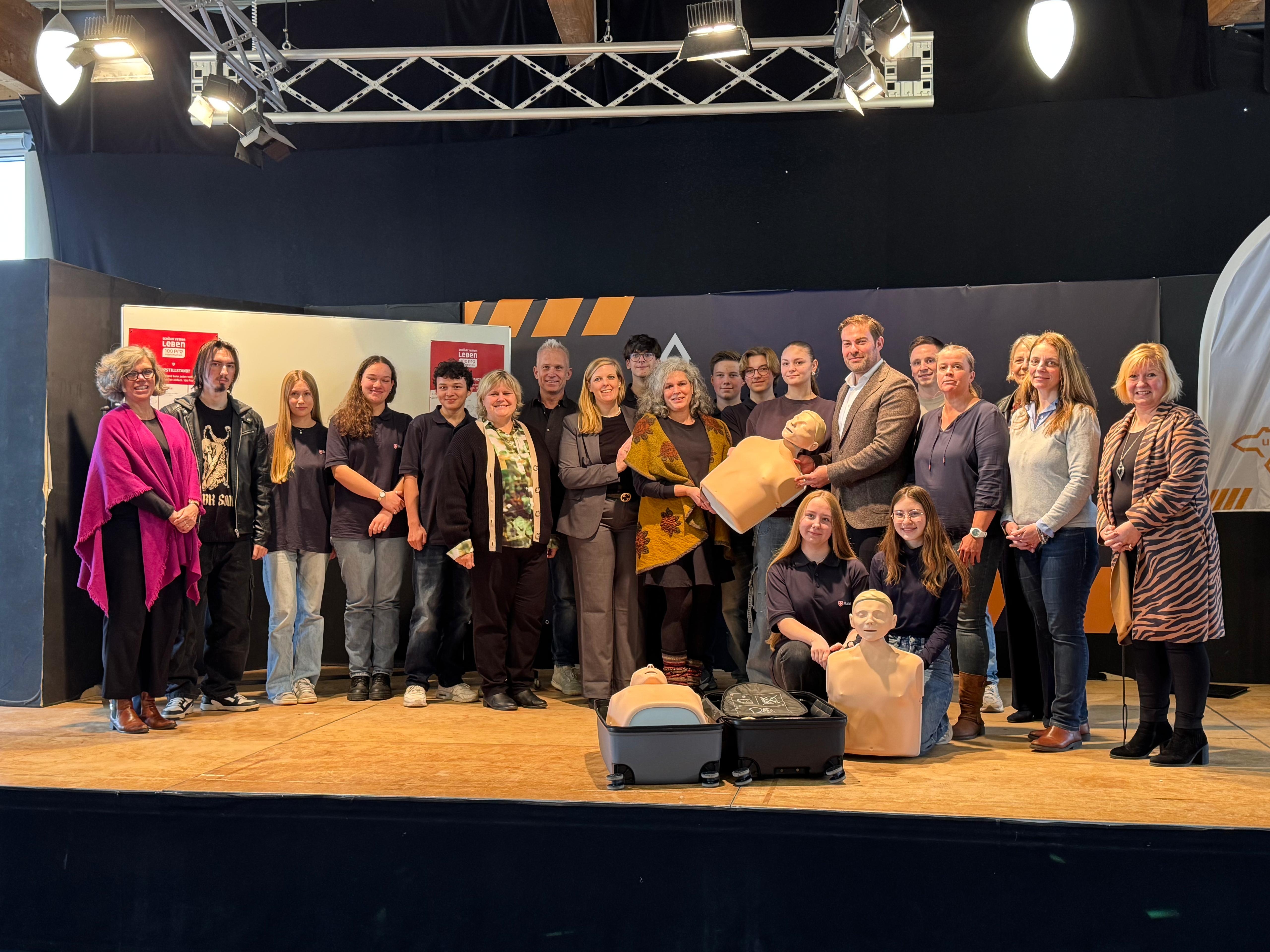 Auf einer Bühne stehen 21 Männer und Frauen mit drei Erste-Hilfe-Puppen Eine davon liegt in einem aufgeklappten Trolley.