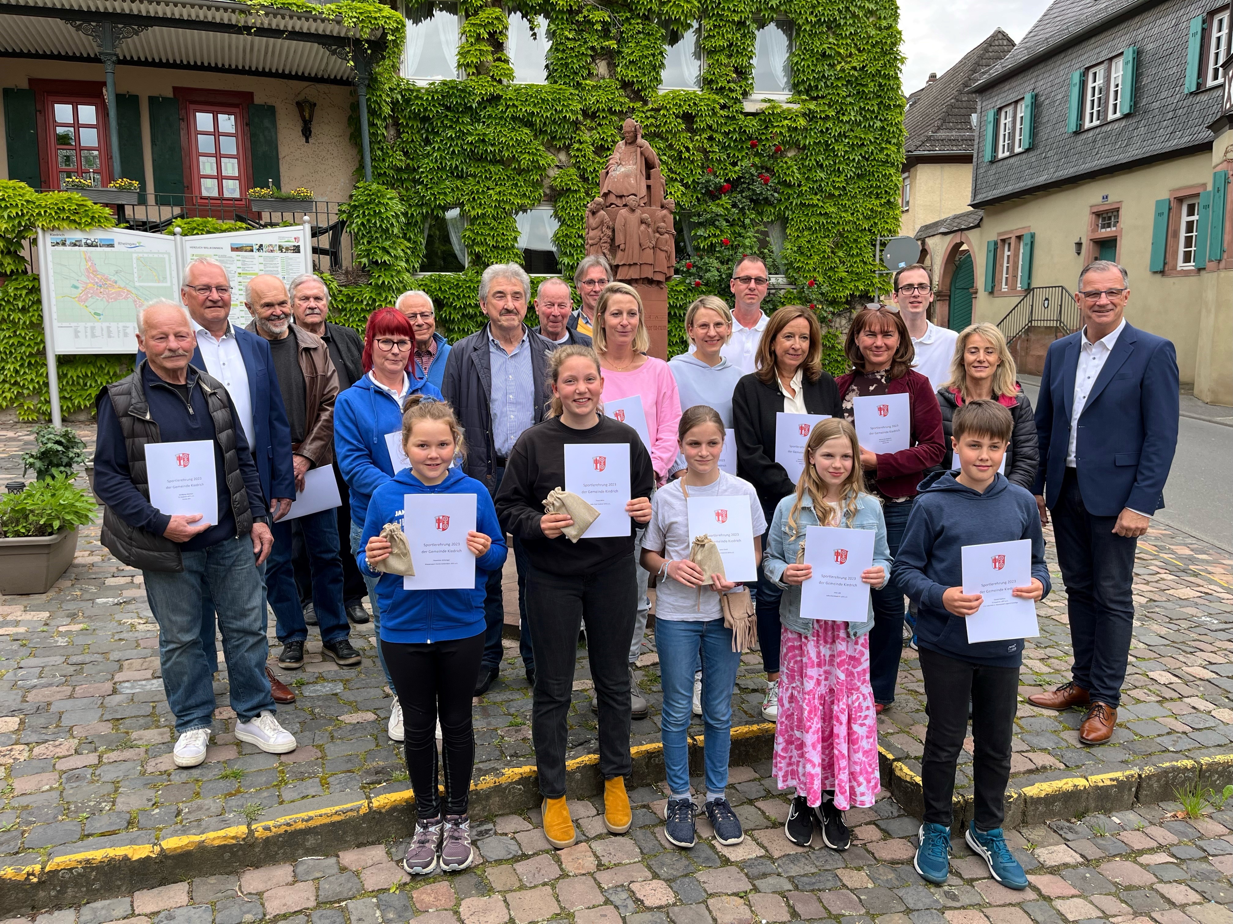 Auf dem Marktplatz mit Pflastersteinen steht eine Gruppe mit Jungen, Mädchen, Frauen und Herren mit Urkunden in der Hand. Herr Bürgermeister Steinmacher steht bei der Gruppe, welche vor einer Werbetafel und einem mit Wein zugewachsenem Haus steht.
