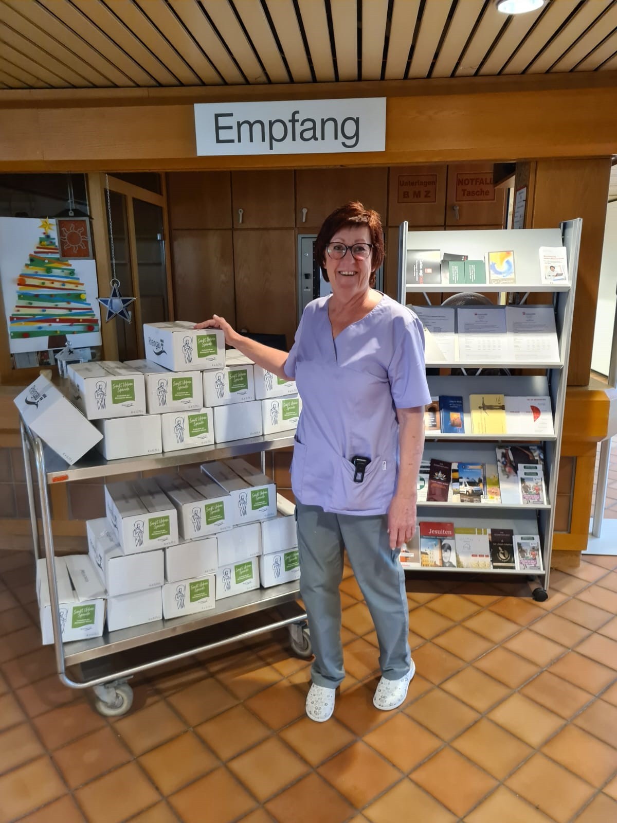 Eine Dame mit Brille und lila Pflege-Shirt und grauer Hose steht neben einem Transportwagen, auf dem 22 Kisten Wein liegen.