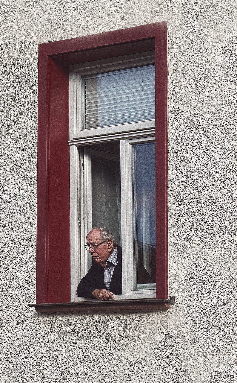 Alter Mann mit Brille schaut aus einem geöffnetem Fenster.