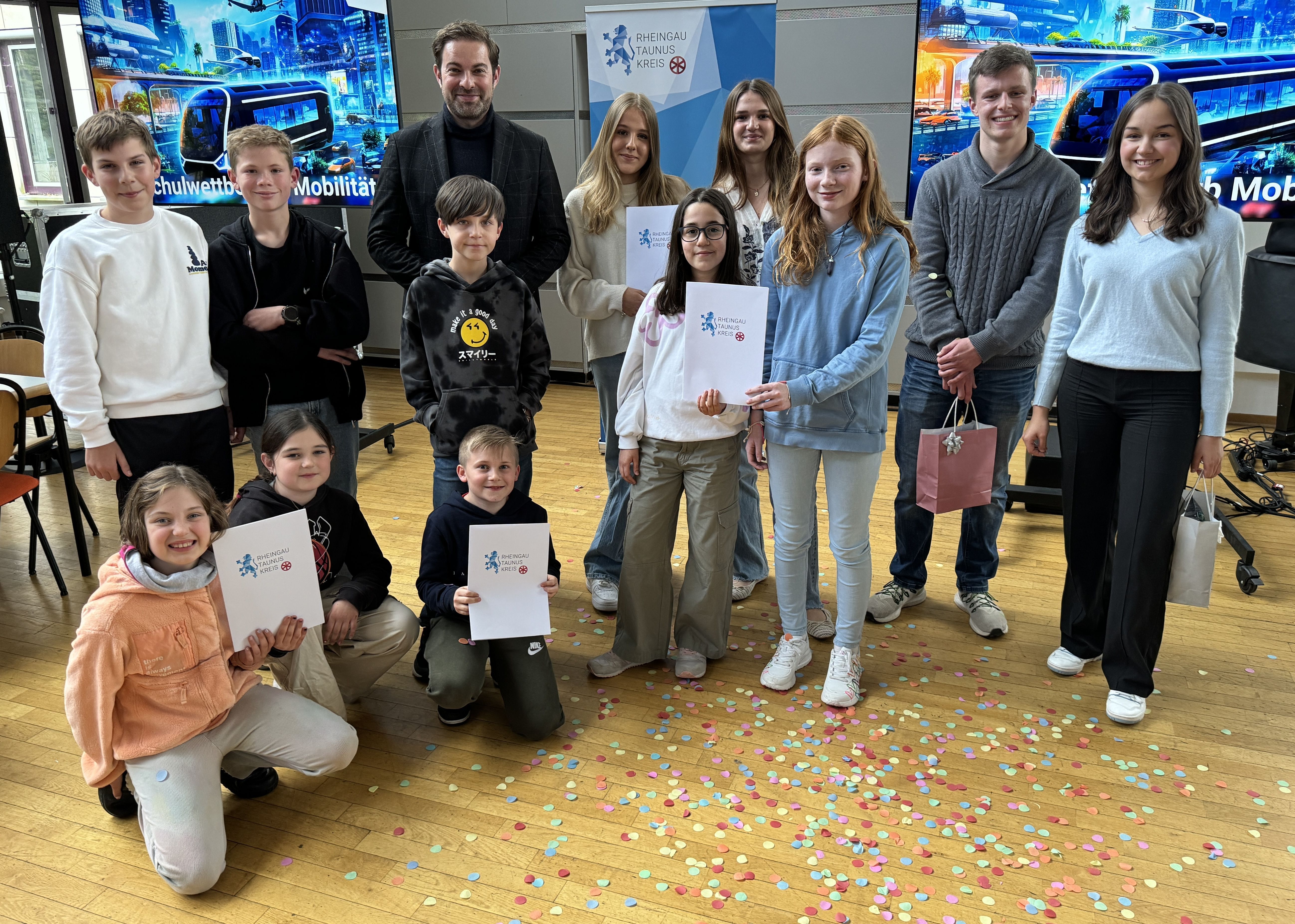 Landrat Sandro Zehner steht mit Schülern, Gewinner und Moderatoren vor dem Banner des Rheingau-Taunus-Kreises und zwei Bildschirmen mit futuristischen Bildern mit Hochhäusern, einer Bahn, Flugelementen und Bäumen. Die Kinder haben zum Teil weiße Urkundenmappen und Geschenktüten in den Händen. Vor der Gruppe liegt Konfetti auf dem Holz-Boden.