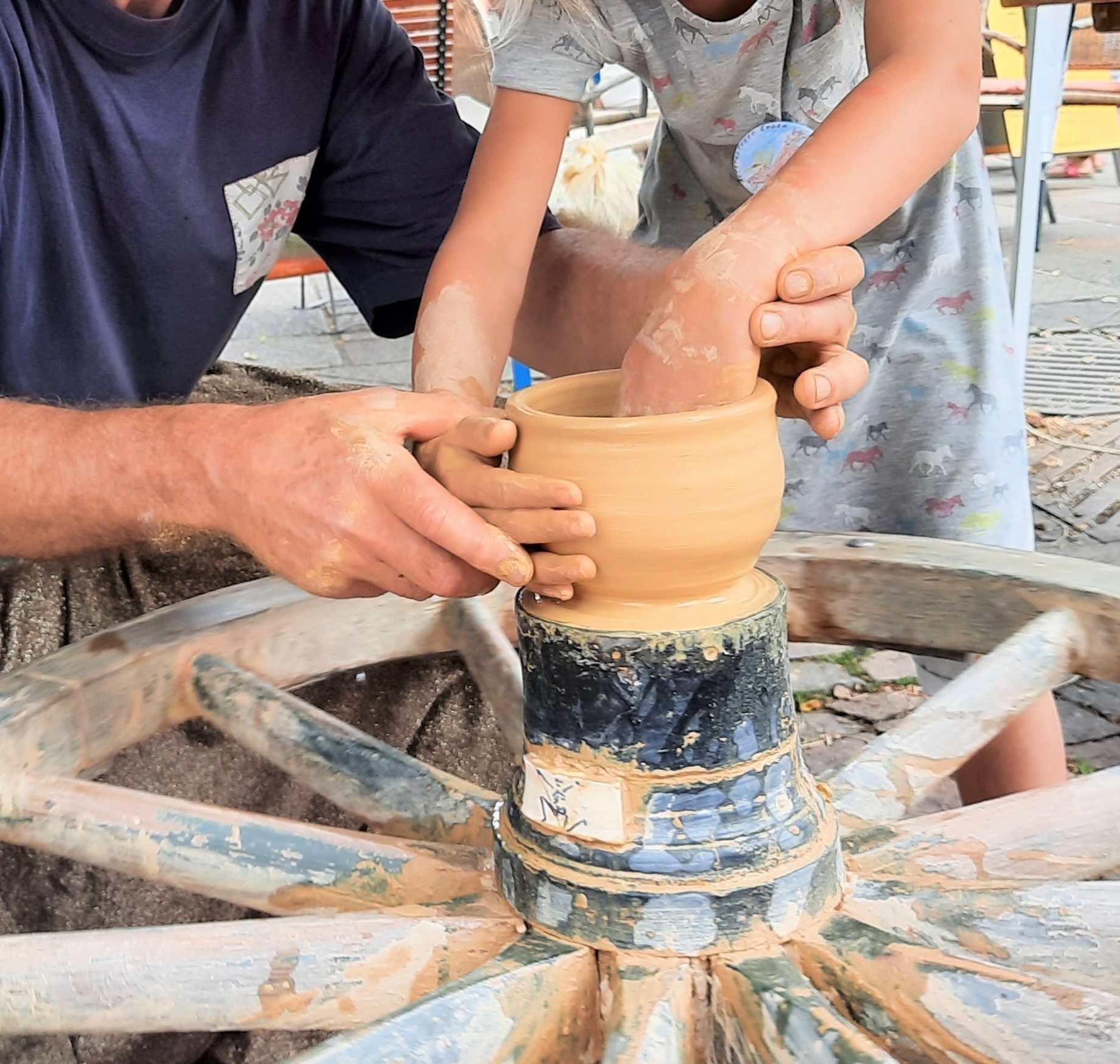 Auf einem Wagenrad aus Holz ist eine Töpfertopf. Zwei Männerhände führen zwei Kinderhände um den Topf zu formen.