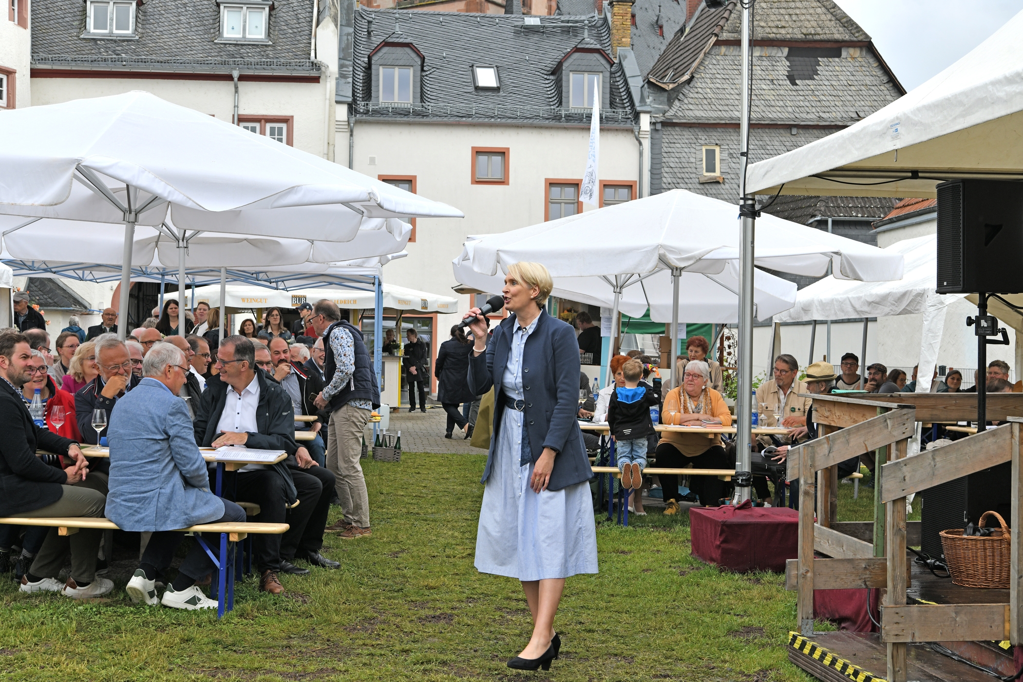 Auf einer Wiese stehen Bierzeltgarnituren und weiße, große, aufgespannte Sonnenschirme. Die Bänke sind besetzt und im Hintergrund ist das Rathaus und ein Weinstand zu sehen. Vorne steht eine Frau mit blonden Haaren, einnem hellbauen Kleid, einer blauen Jacke und blauen Pumps. Sie hält ein Mikrofon in der Hand und singt.